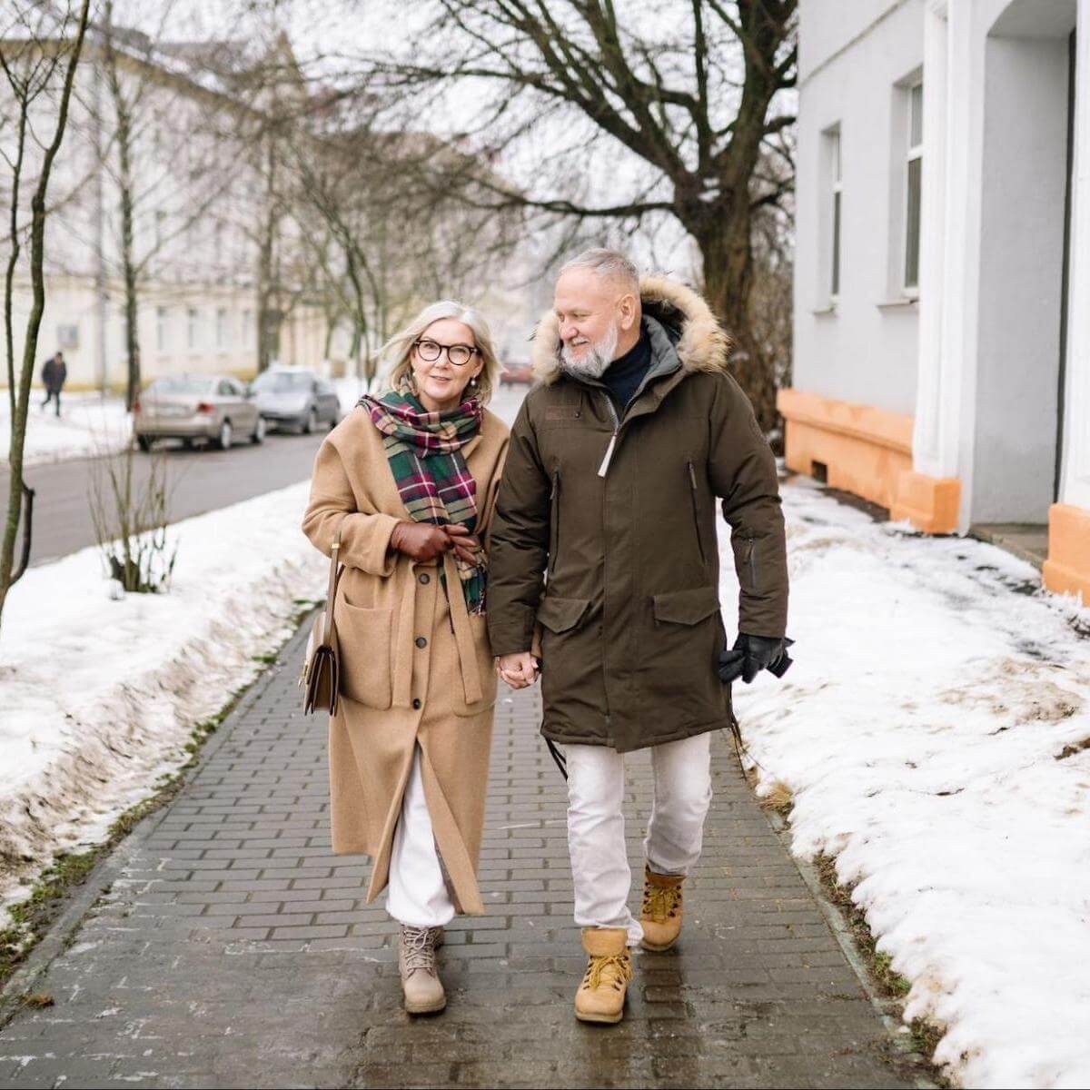 Elderly couple holding hands walking on snowy sidewalk in winter coats with bare trees in residential street