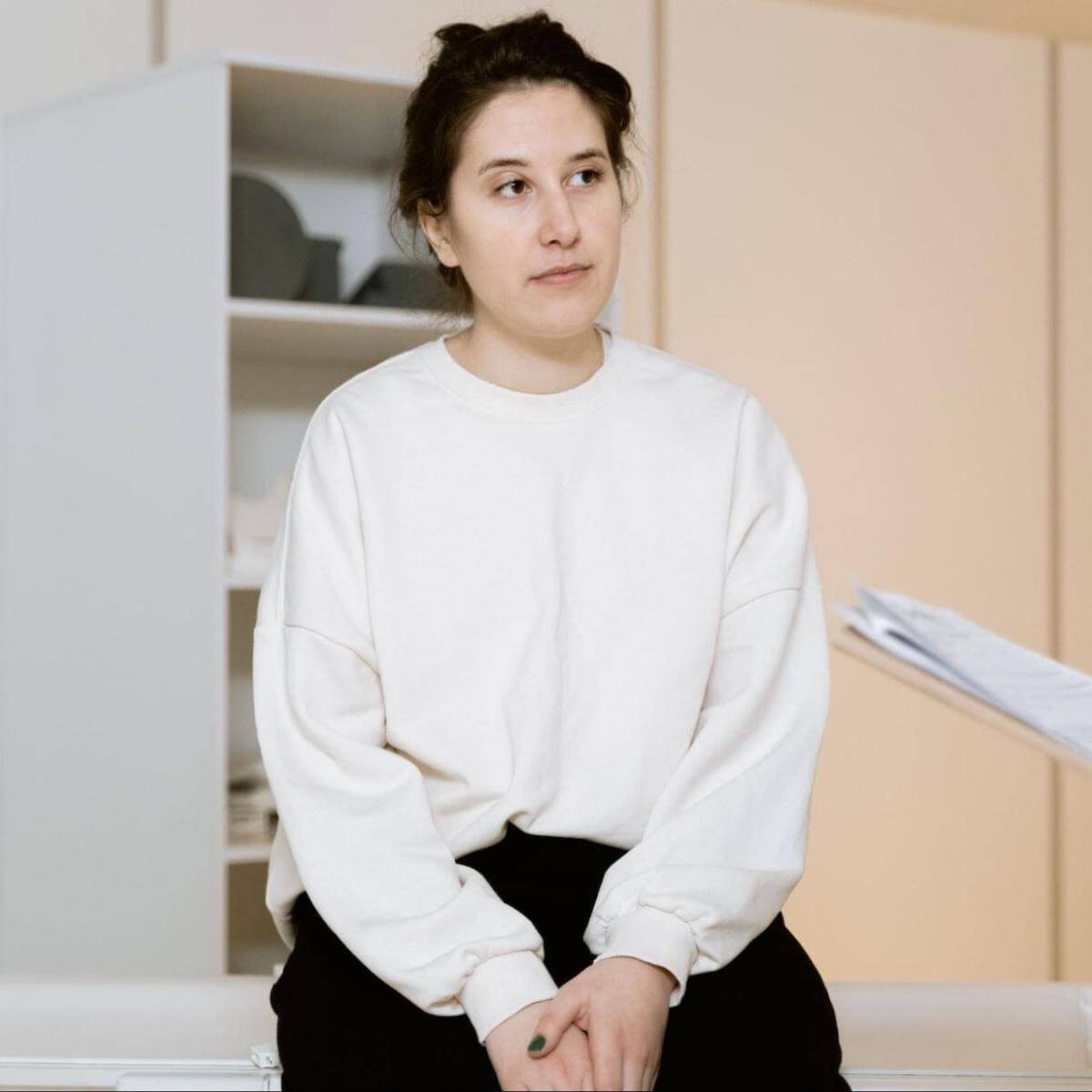 Young woman in white sweatshirt and black pants sitting in minimal interior space with thoughtful expression