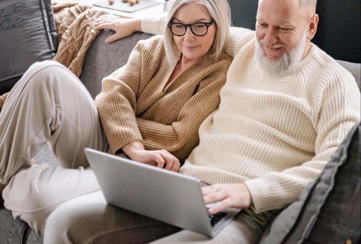 Middle-aged couple smiling while viewing a laptop together on a gray couch with blankets