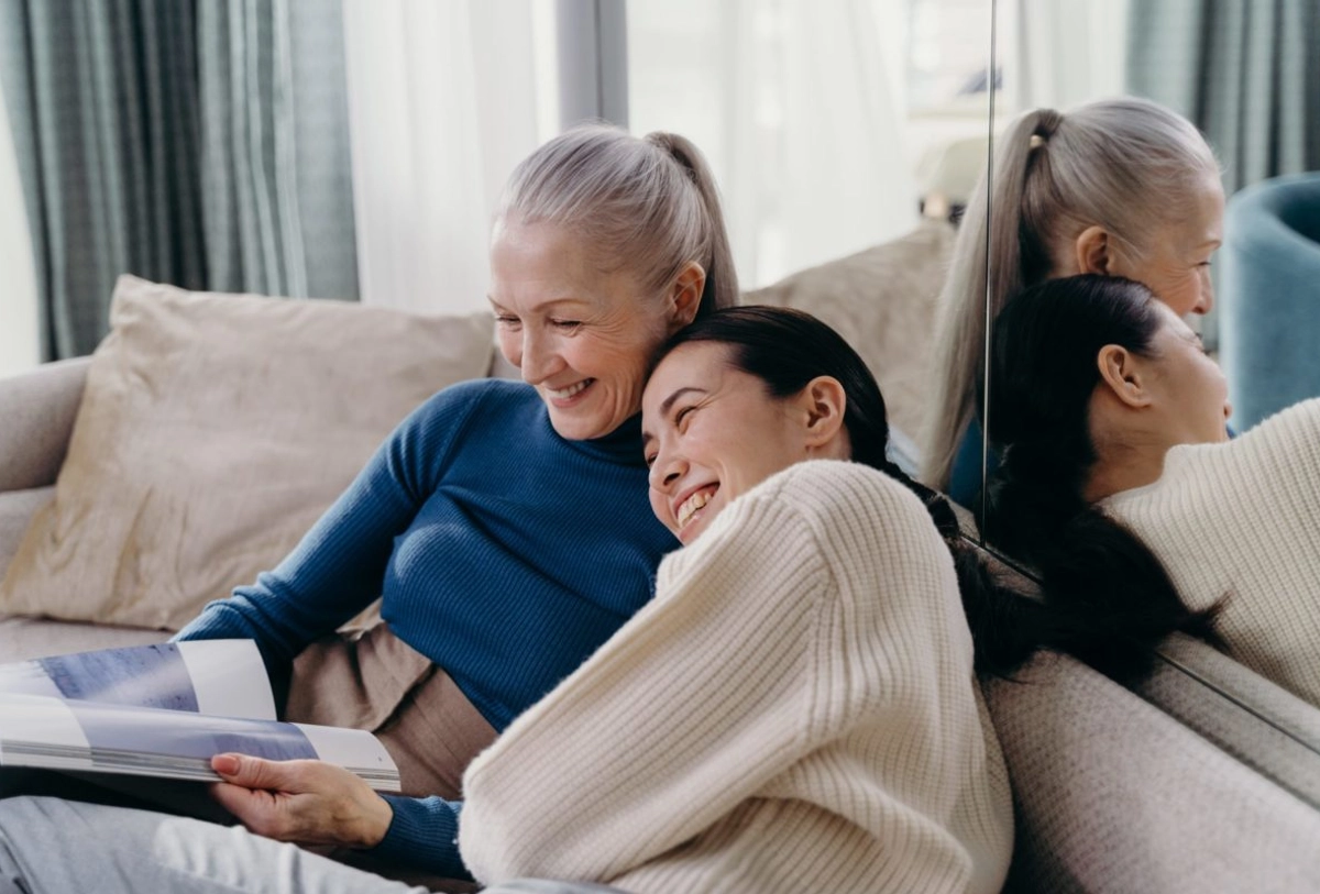 Older and younger woman smiling together on couch looking at laptop with curtains and pillows in modern living room