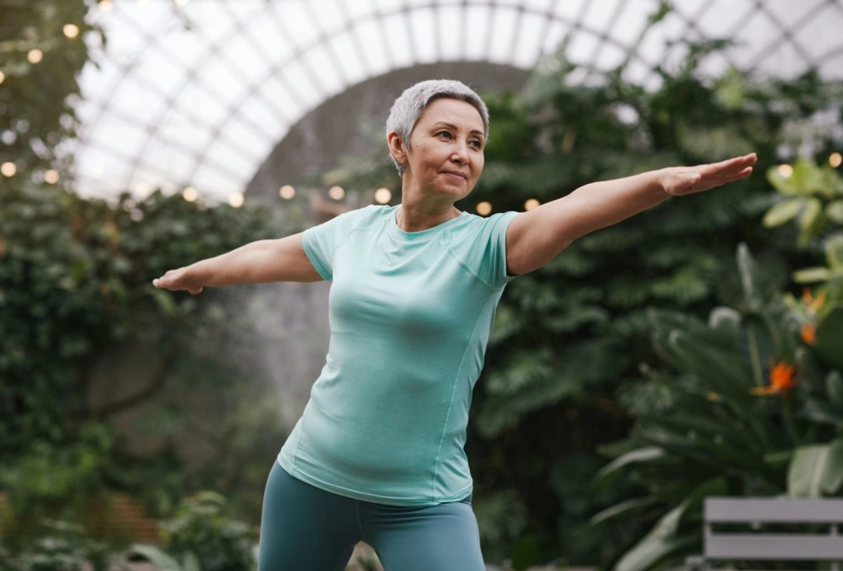 Mature woman in turquoise athletic wear performing balance pose with arms extended in greenhouse with plants