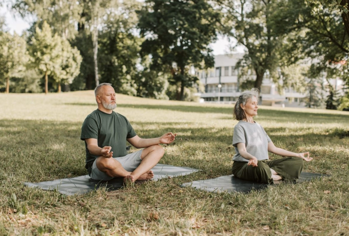 Mature man and woman meditating on yoga mats outdoors in park with trees and building in background