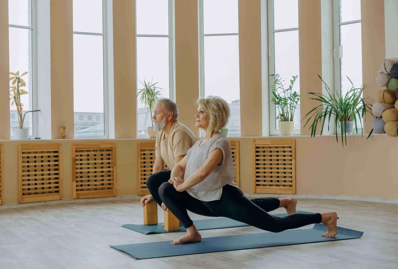 Two mature adults performing yoga stretches on mats in bright studio with floor-to-ceiling windows and plants