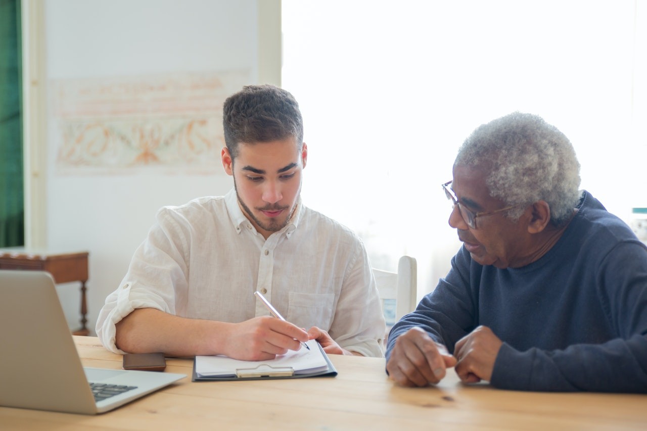 Young man and elderly man reviewing documents together at wooden table with laptop