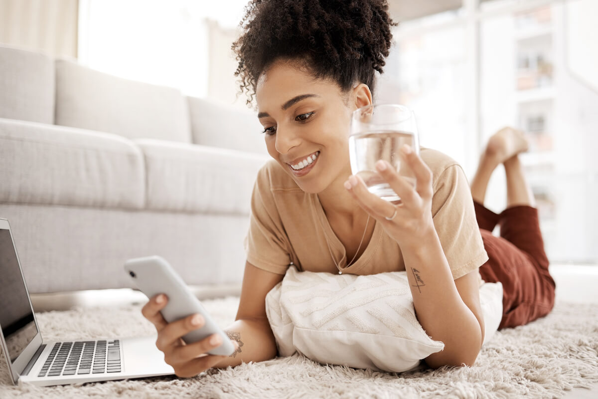 Smiling woman lying on carpet at home, holding phone and water glass while working on laptop