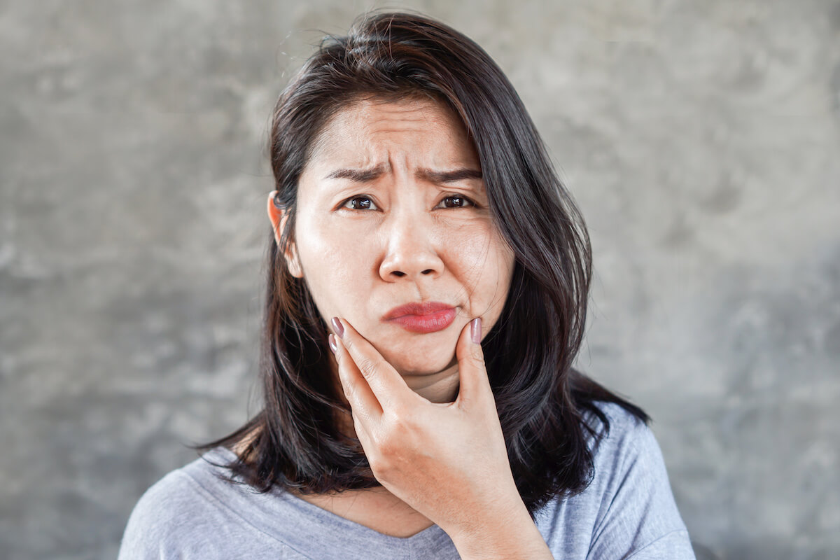 Woman holding her cheek with pained expression against gray concrete background