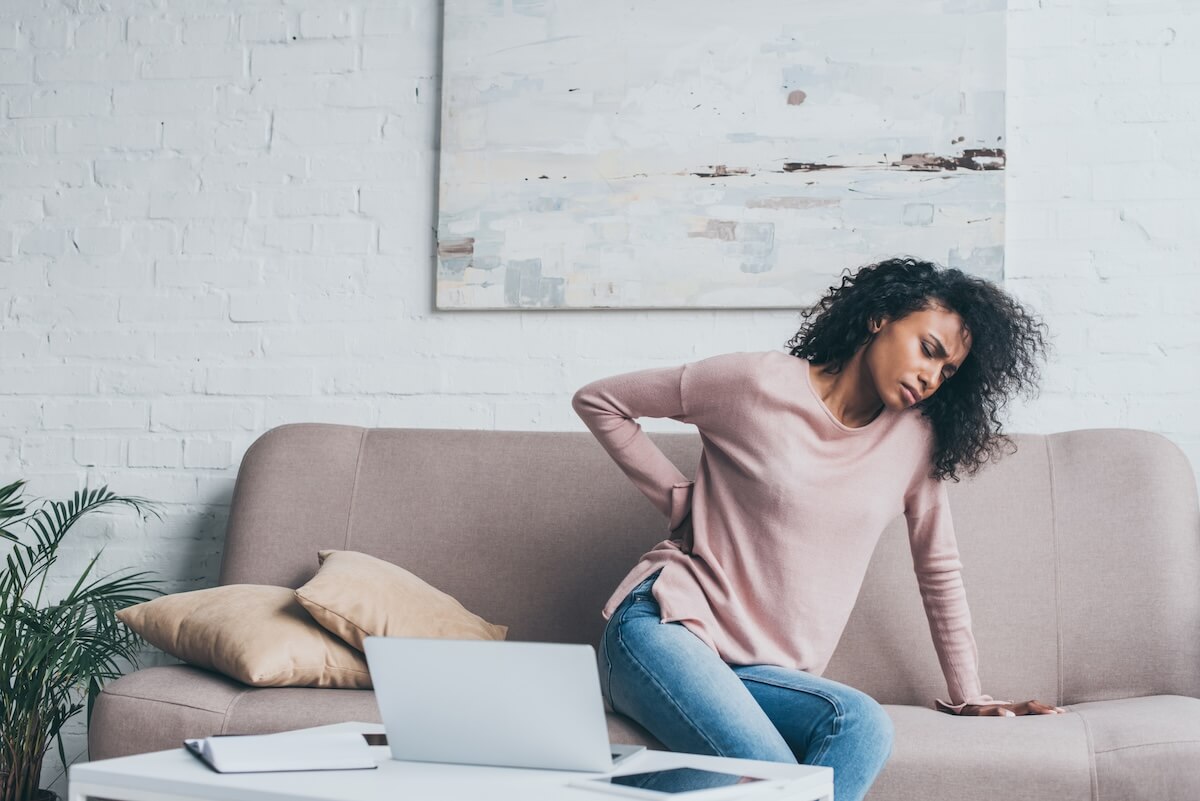 Woman sitting on couch holding her lower back in pain while working on laptop at home