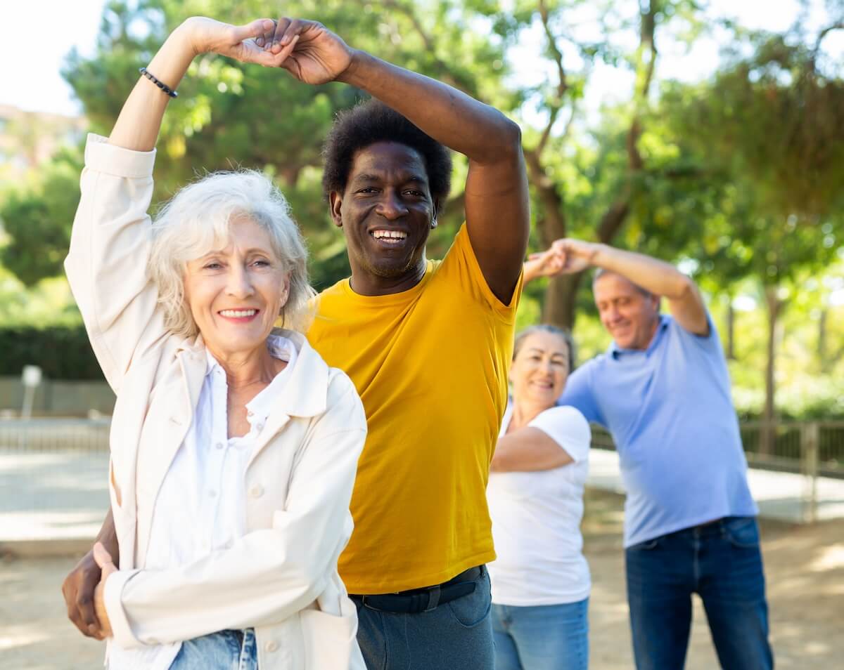 Group of diverse adults exercising outdoors with raised arms in dance-like movements against green park background