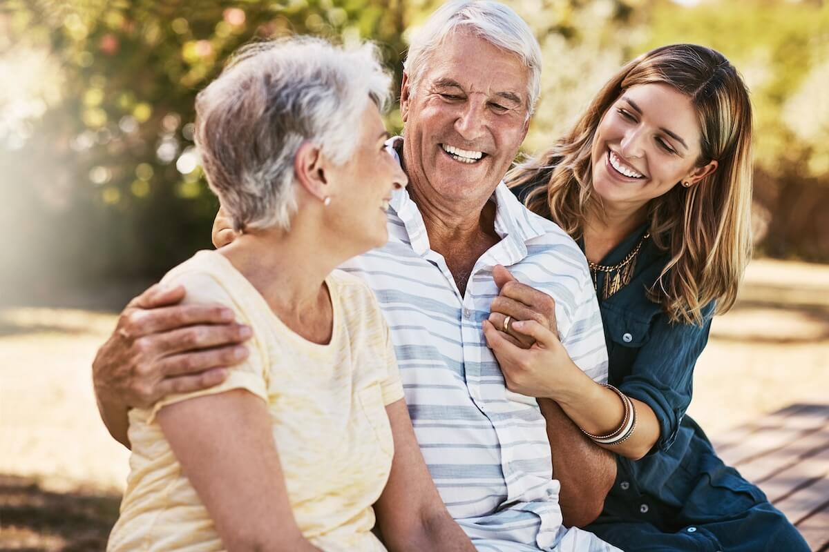 Three generations sharing joyful moment outdoors, senior couple with younger woman laughing in sunlit natural setting