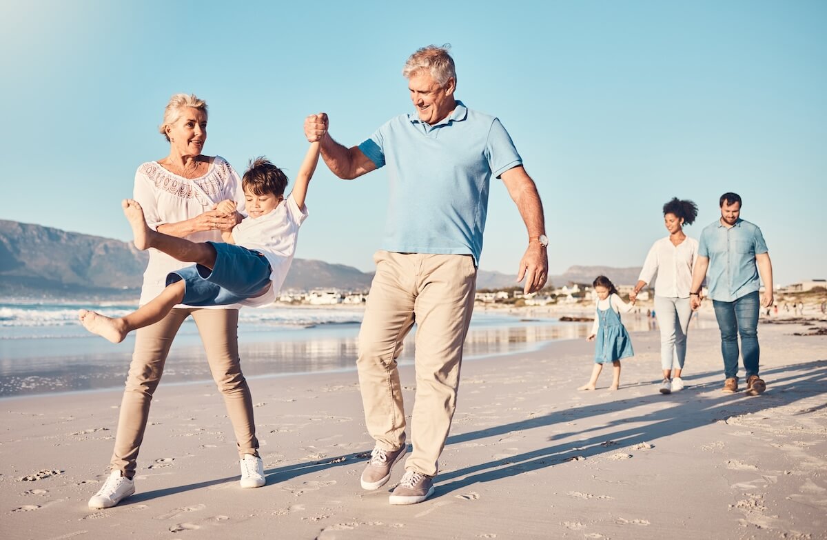 Multigenerational family enjoying beach walk, grandparents swinging child while parents follow with daughter, mountains behind