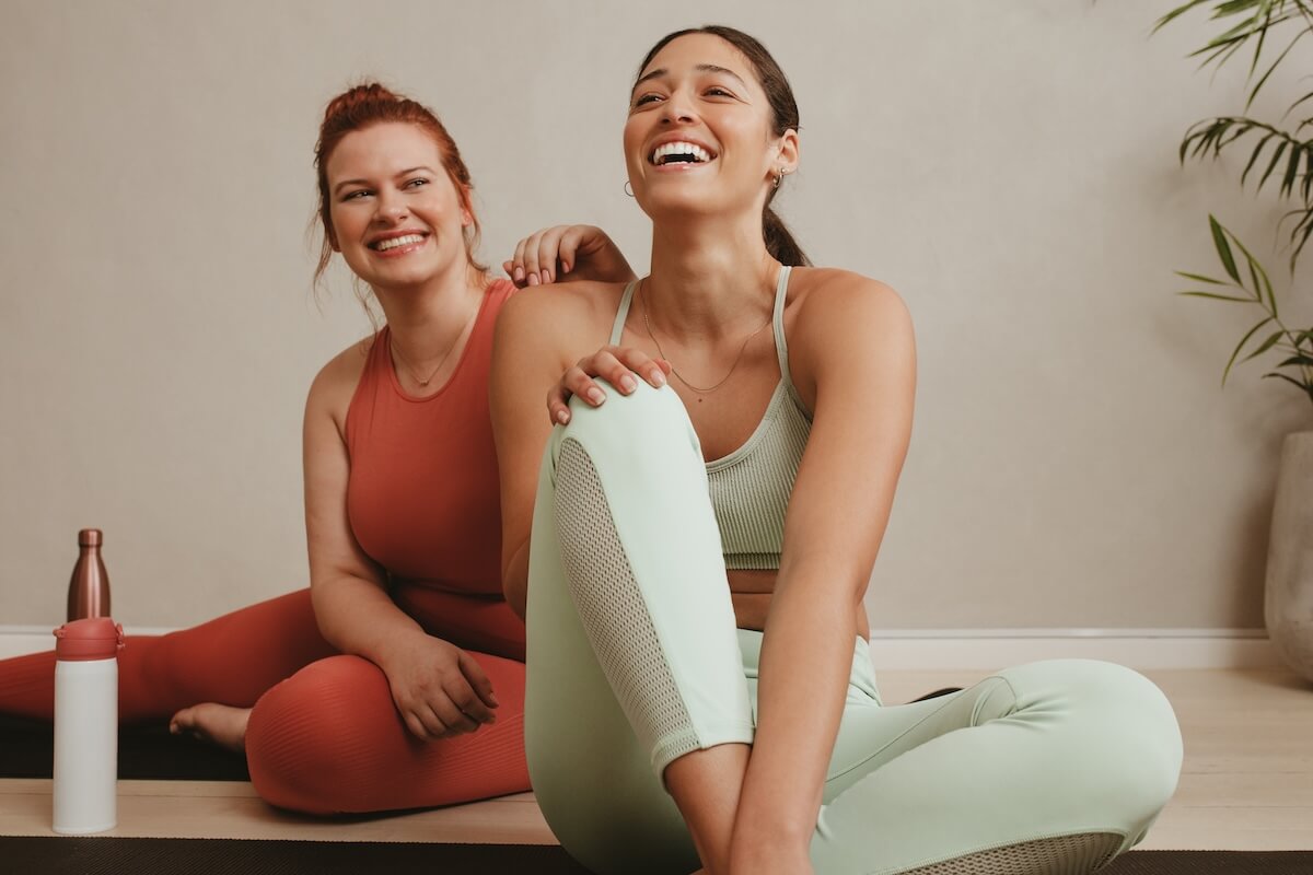 Two friends in workout attire laughing together on yoga mats, wearing coral and mint exercise sets