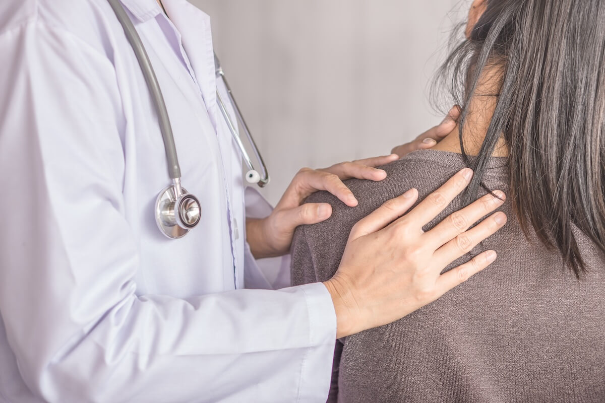 Healthcare provider in white coat with stethoscope examining patient's shoulder, hands placed supportively on upper back