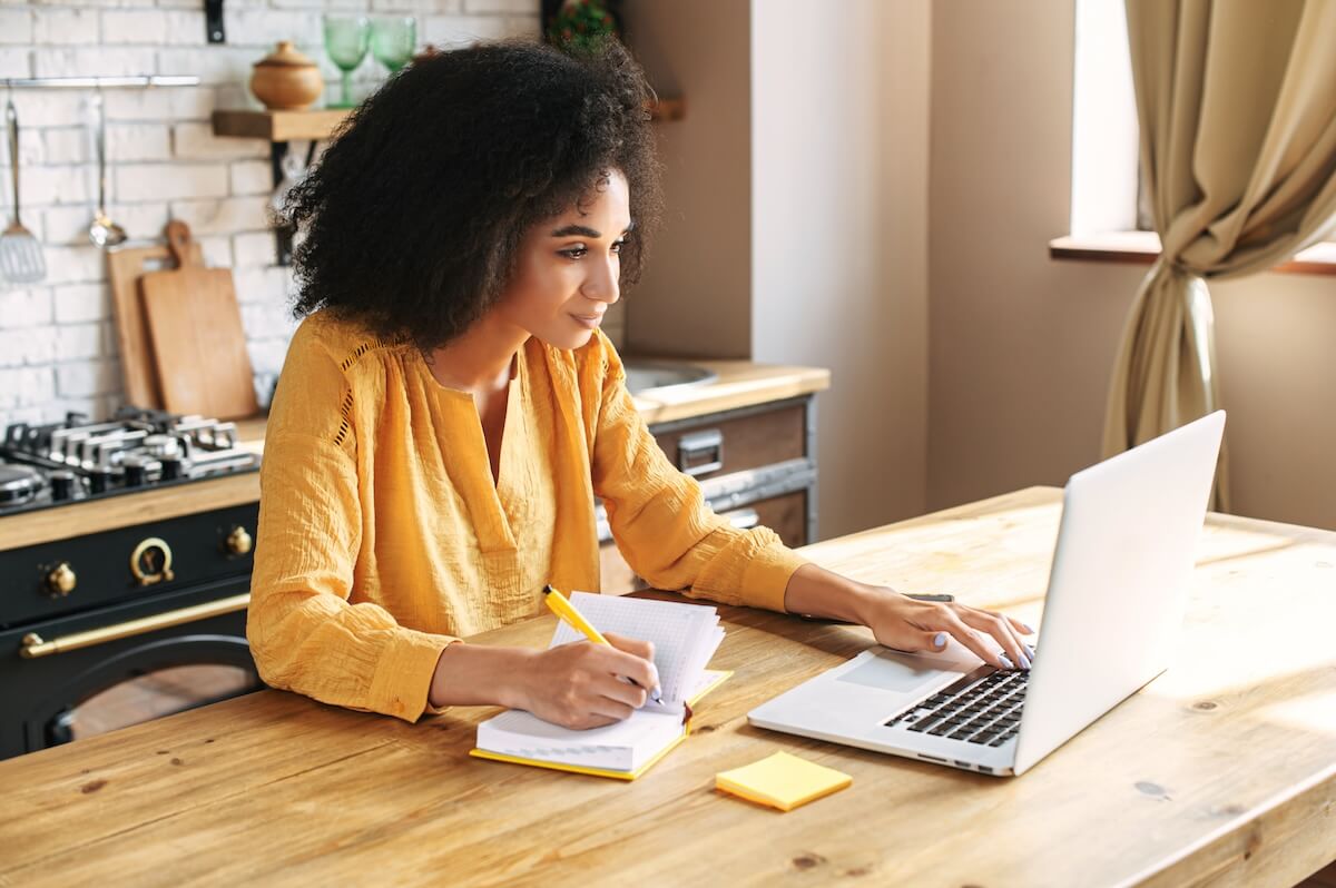 Woman in yellow shirt taking notes while working on laptop at wooden kitchen table