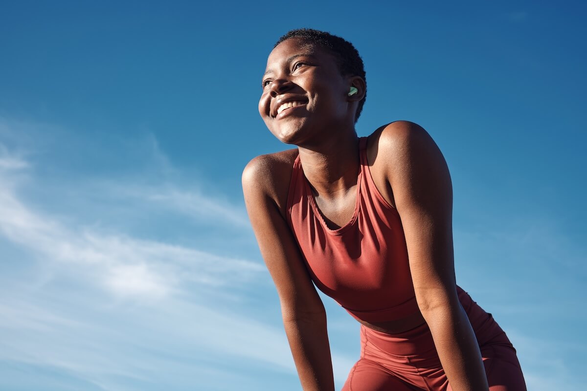 Person in terracotta athletic wear smiling against bright blue sky, wearing green earbuds during outdoor activity