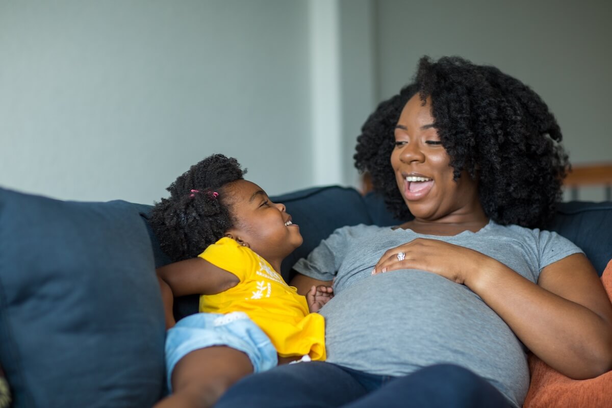 Pregnant woman in gray shirt and child in yellow top sharing joyful moment on blue couch, both laughing together