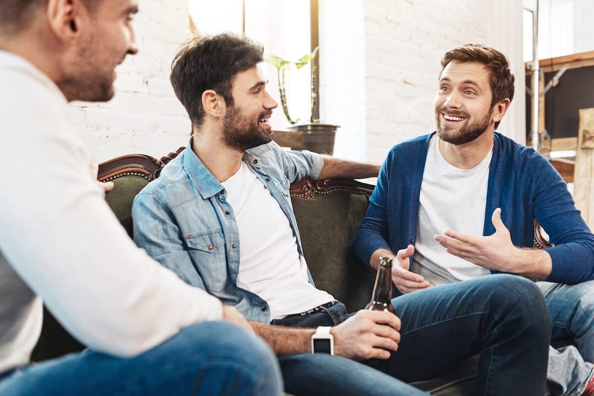 Three people conversing on a couch in casual setting with white brick walls, sharing drinks and laughing during social gathering