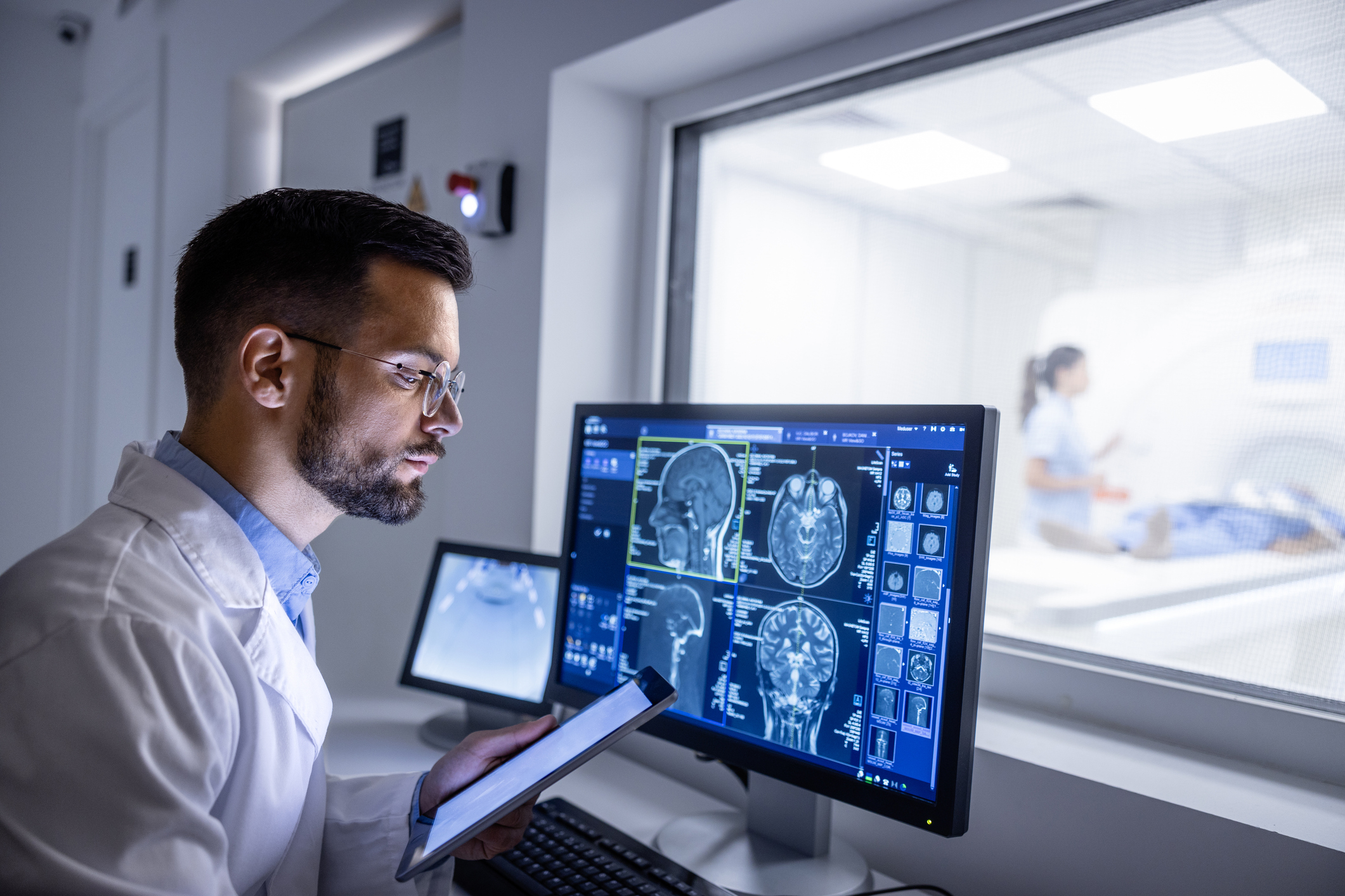 Medical professional viewing brain MRI scans on monitor, tablet in hand, imaging room visible through window