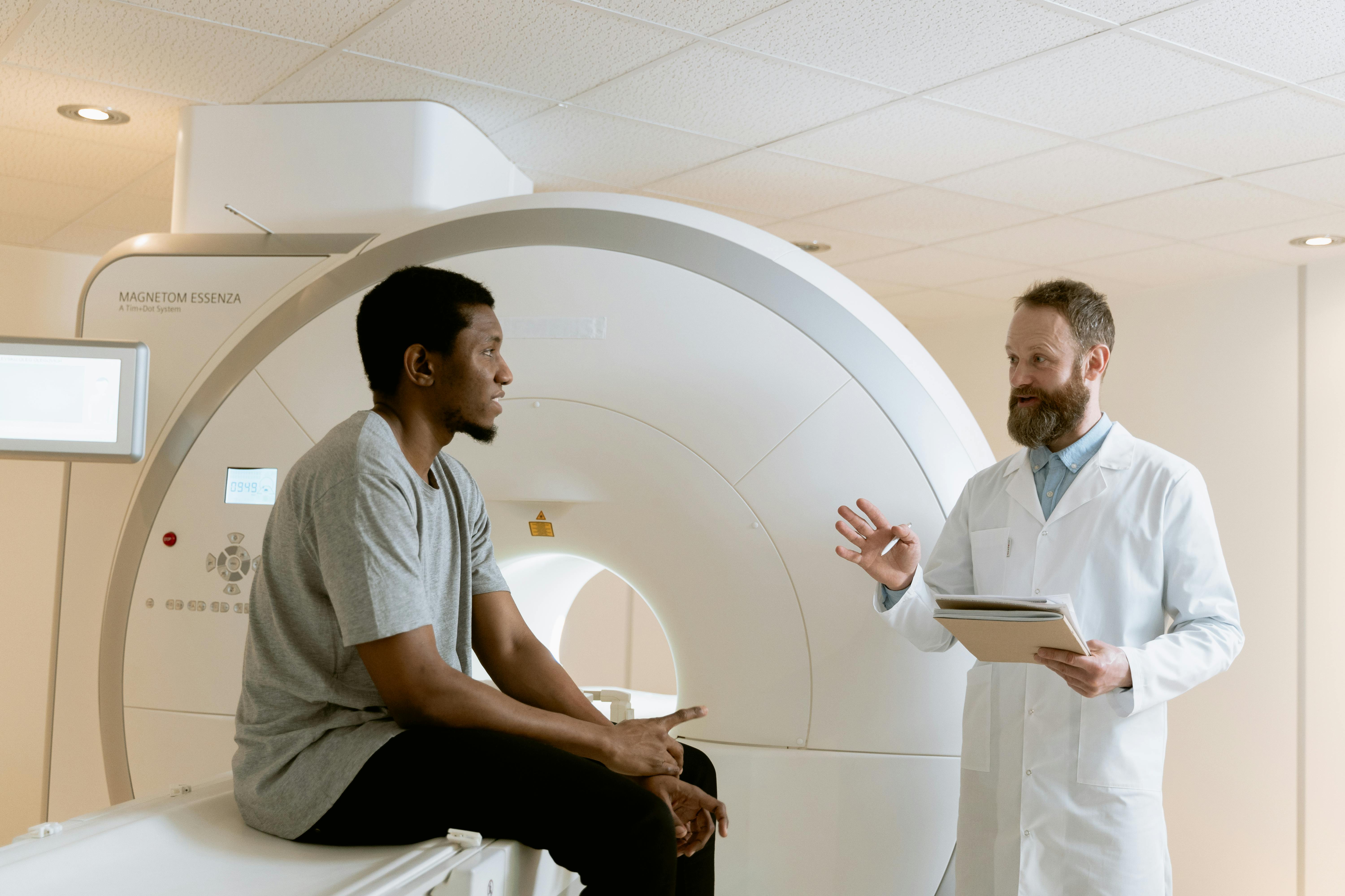 Doctor in white coat consulting with patient seated on MRI machine while holding tablet