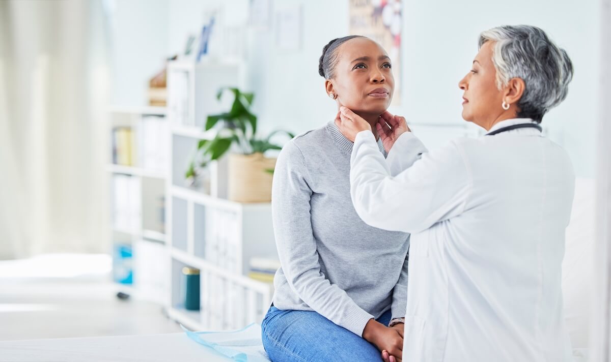 Doctor in white coat examining patient's throat, patient in gray sweater sitting on exam table in bright office