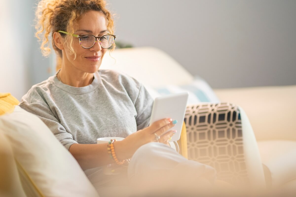 Person with curly hair and glasses in gray sweatshirt using tablet on couch, relaxed home