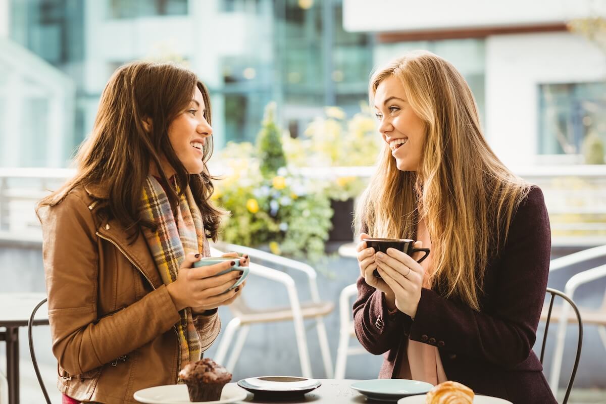 Two women smiling while holding phones at outdoor café table with city backdrop