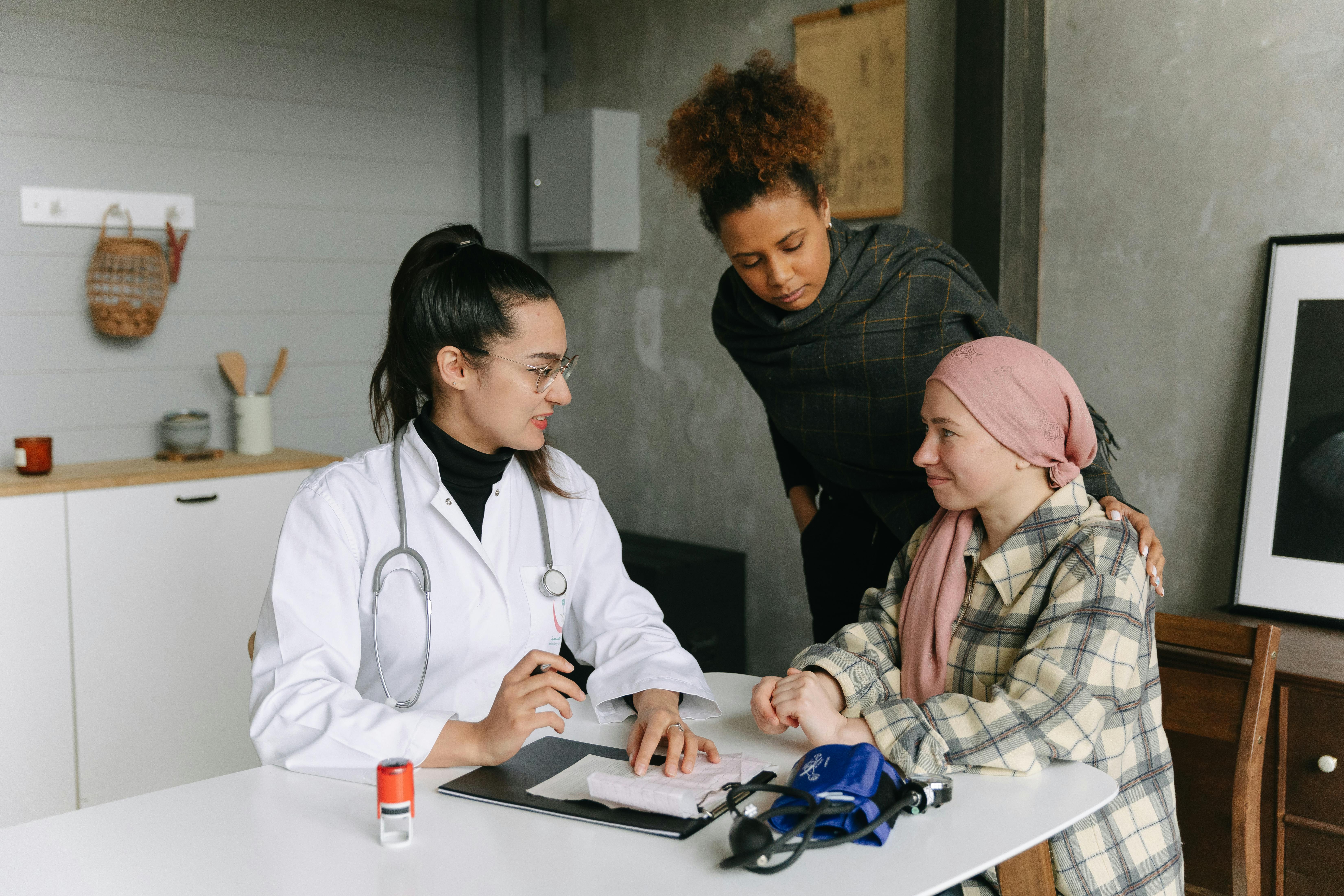 Female doctor consulting with patient wearing pink headscarf and companion while reviewing tablet at clinic