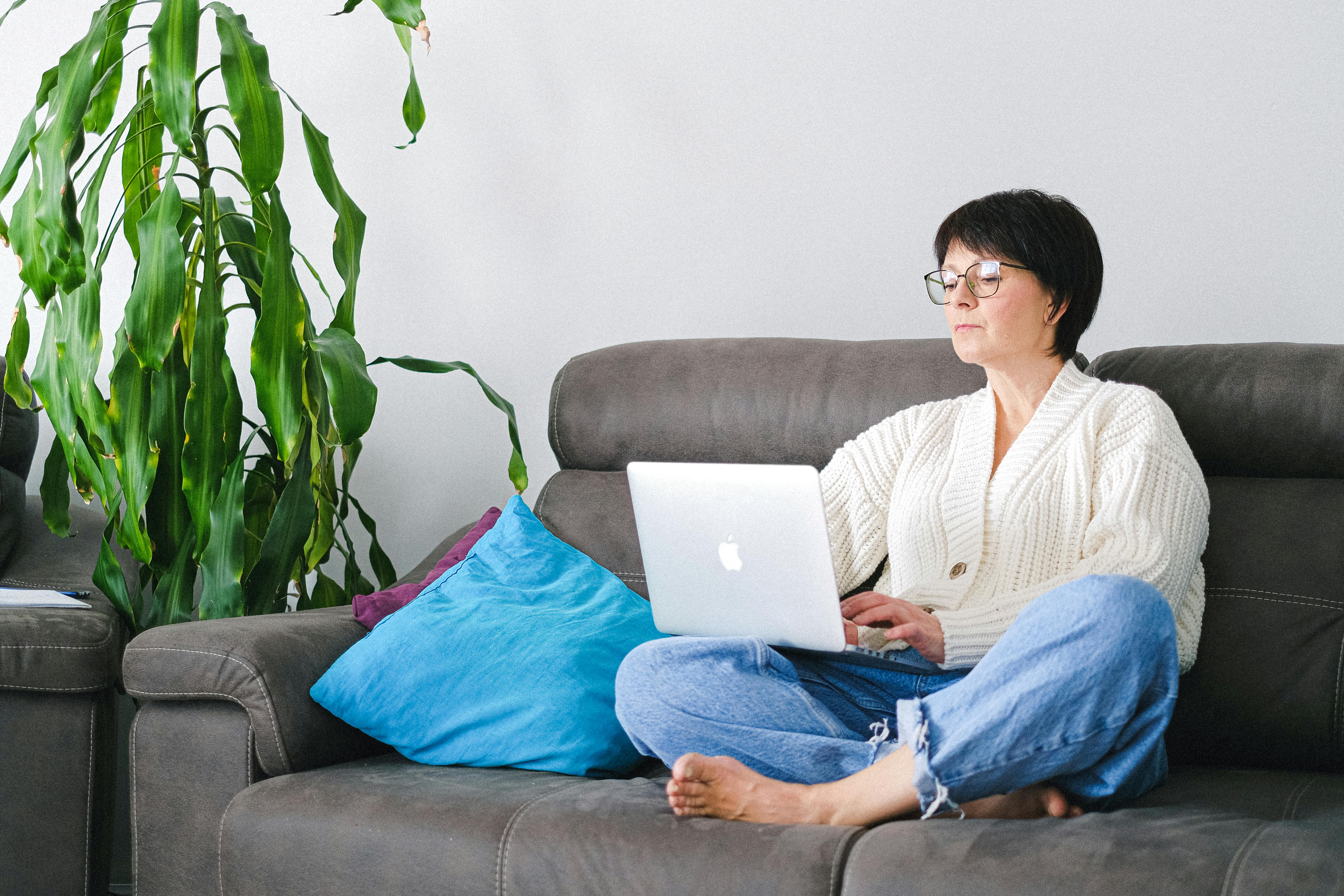 Woman on gray couch working on laptop with blue pillow and potted green plant beside her