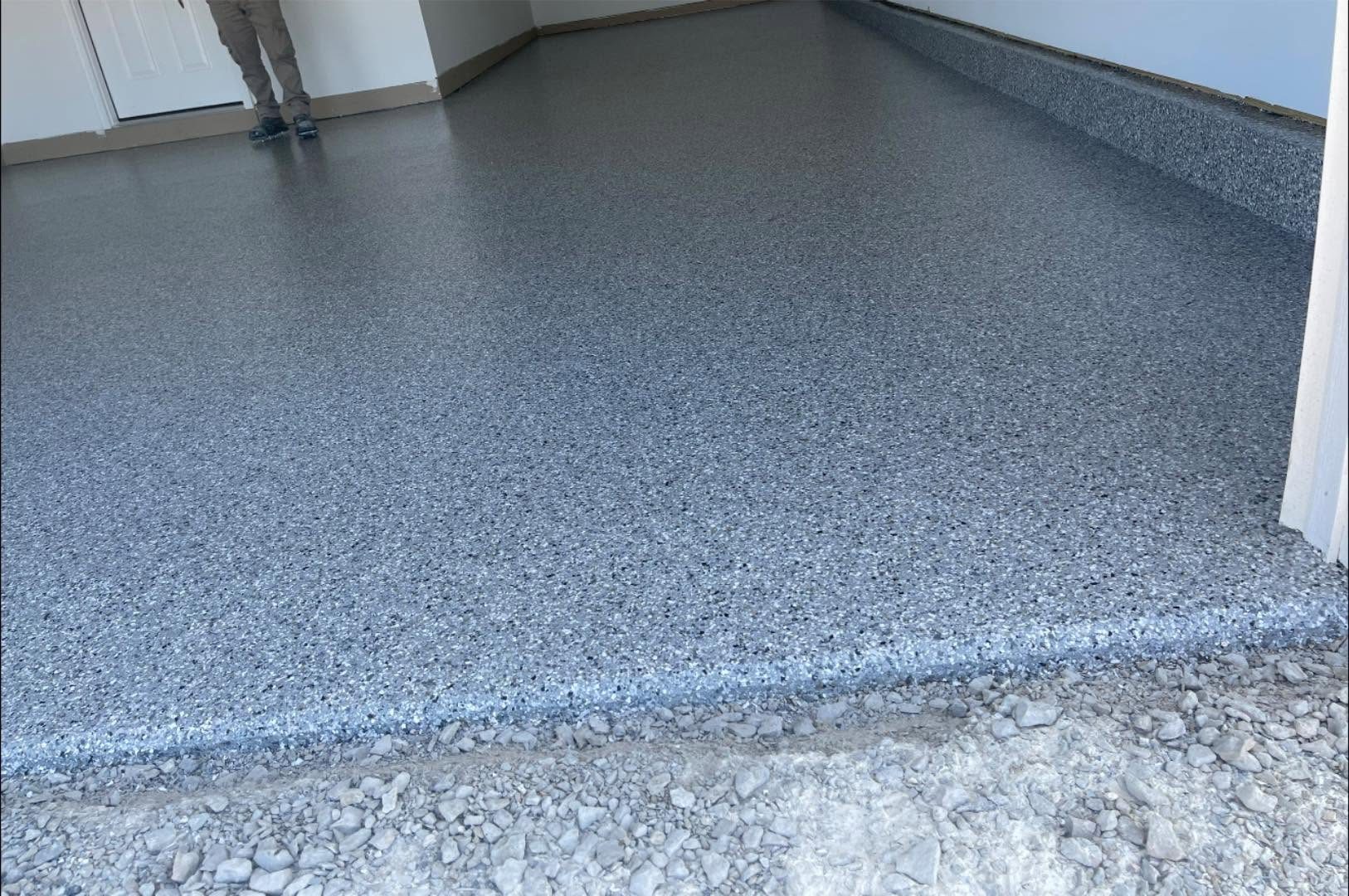 Garage floor with a fresh, speckled epoxy flake coating and a rough gravel driveway outside.