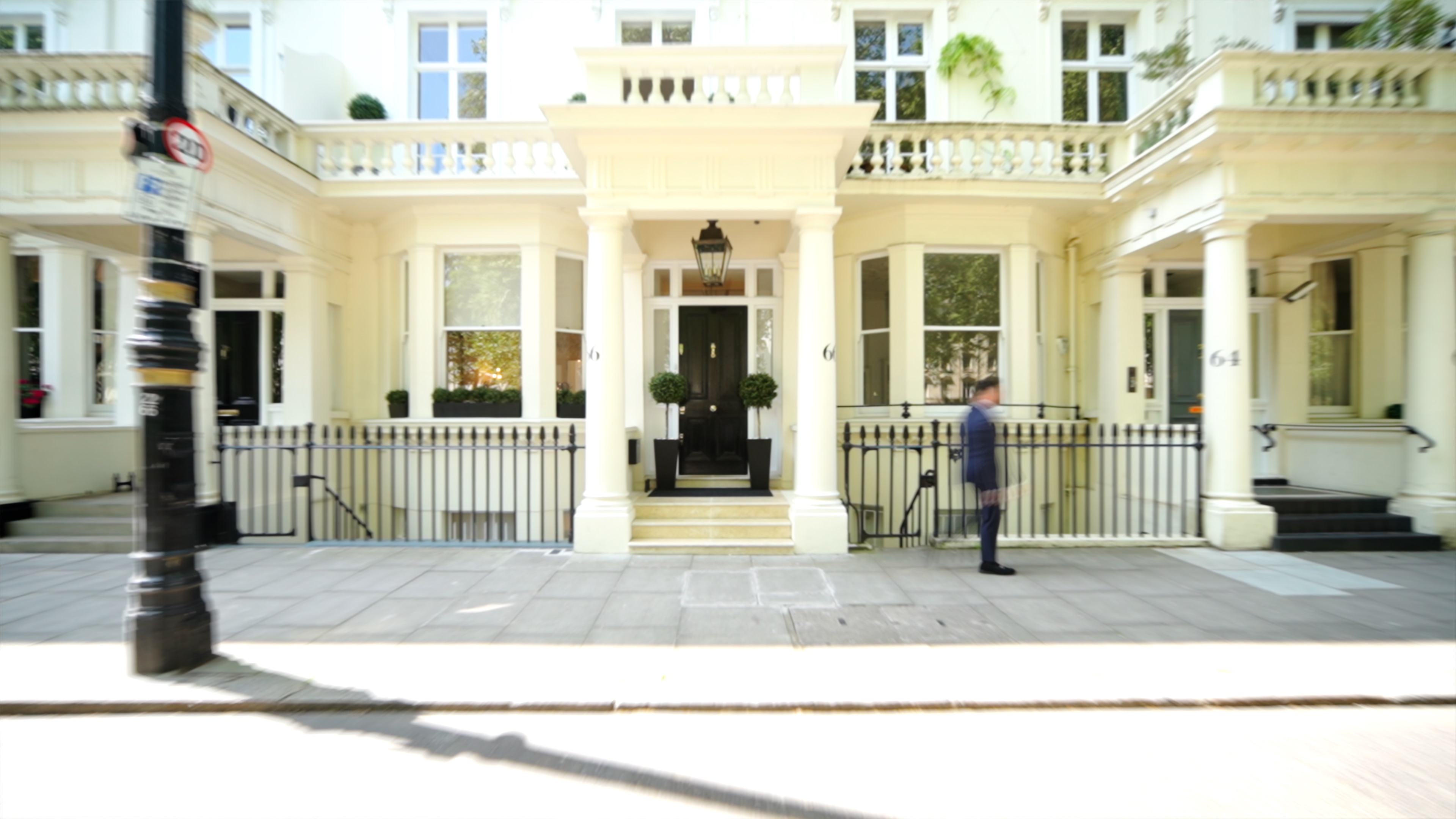 Light-colored building with black front door, columns, and a person blurred walking on the sidewalk.