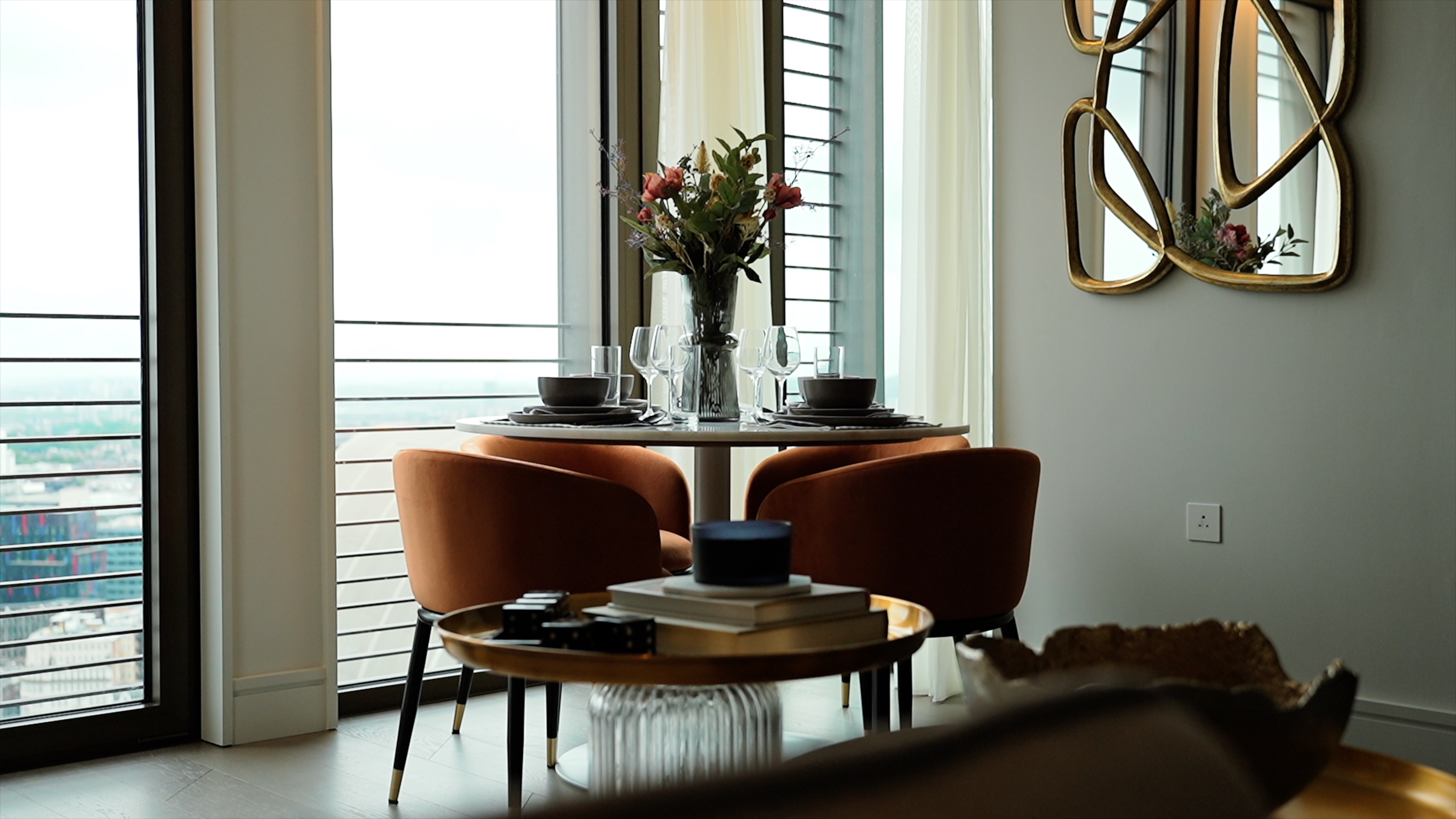 Modern dining area with round table, four brown chairs, flower vase, and city view windows.