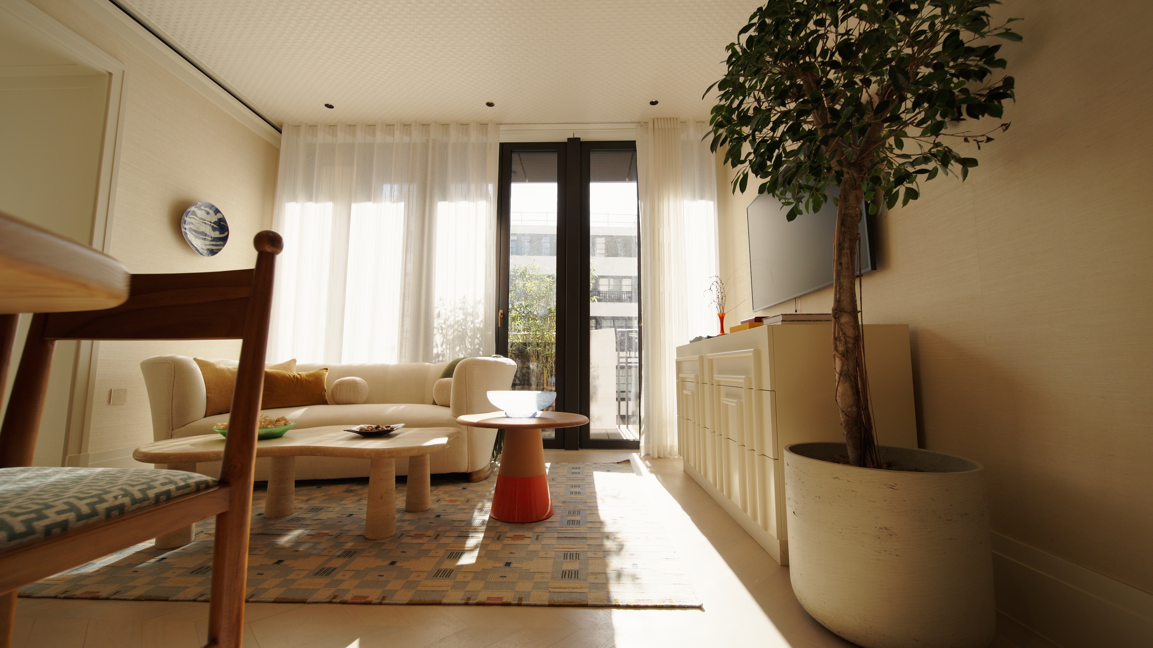 Bright living room with a beige sofa, wooden tables, large plant, and sheer curtains over glass doors.