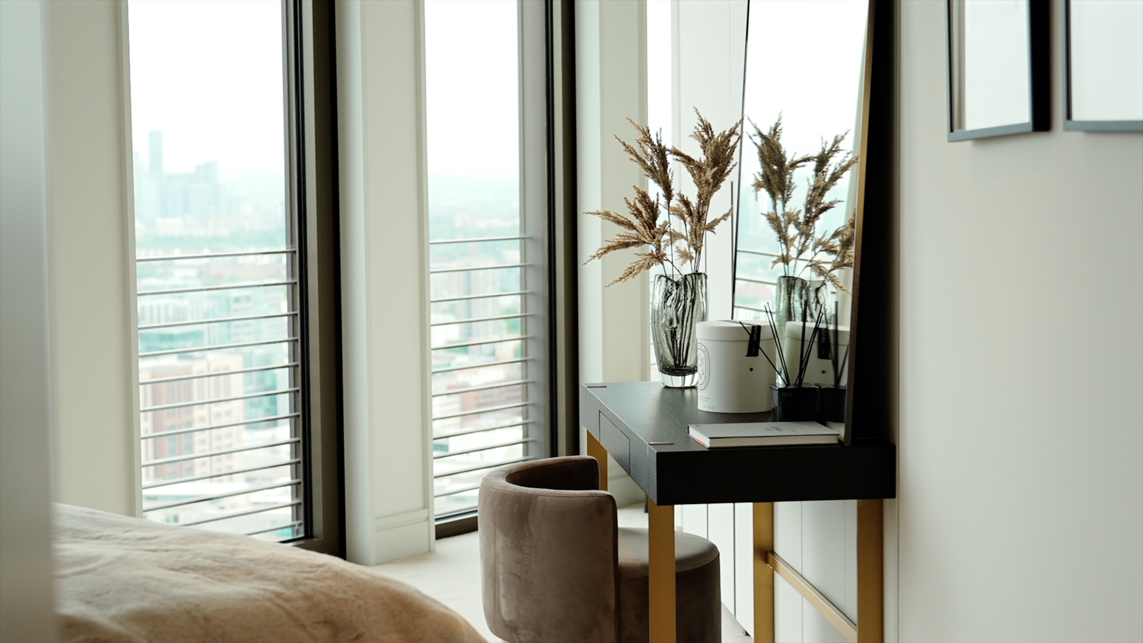 Modern dressing table with vase, book, and reed diffuser beside a round chair near large windows.