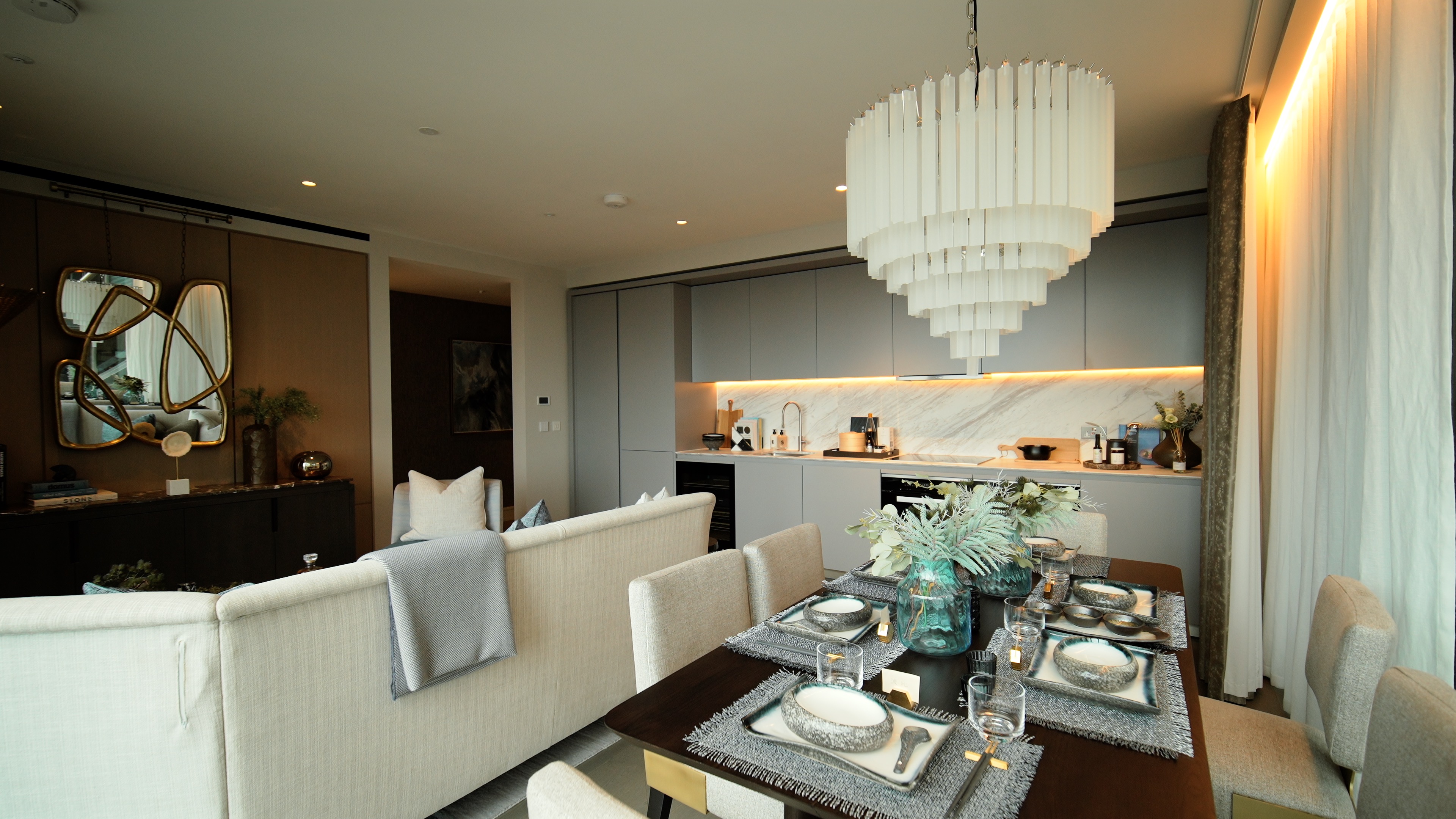 Modern dining and living area with set table, beige couch, and gray kitchen cabinets under chandelier.