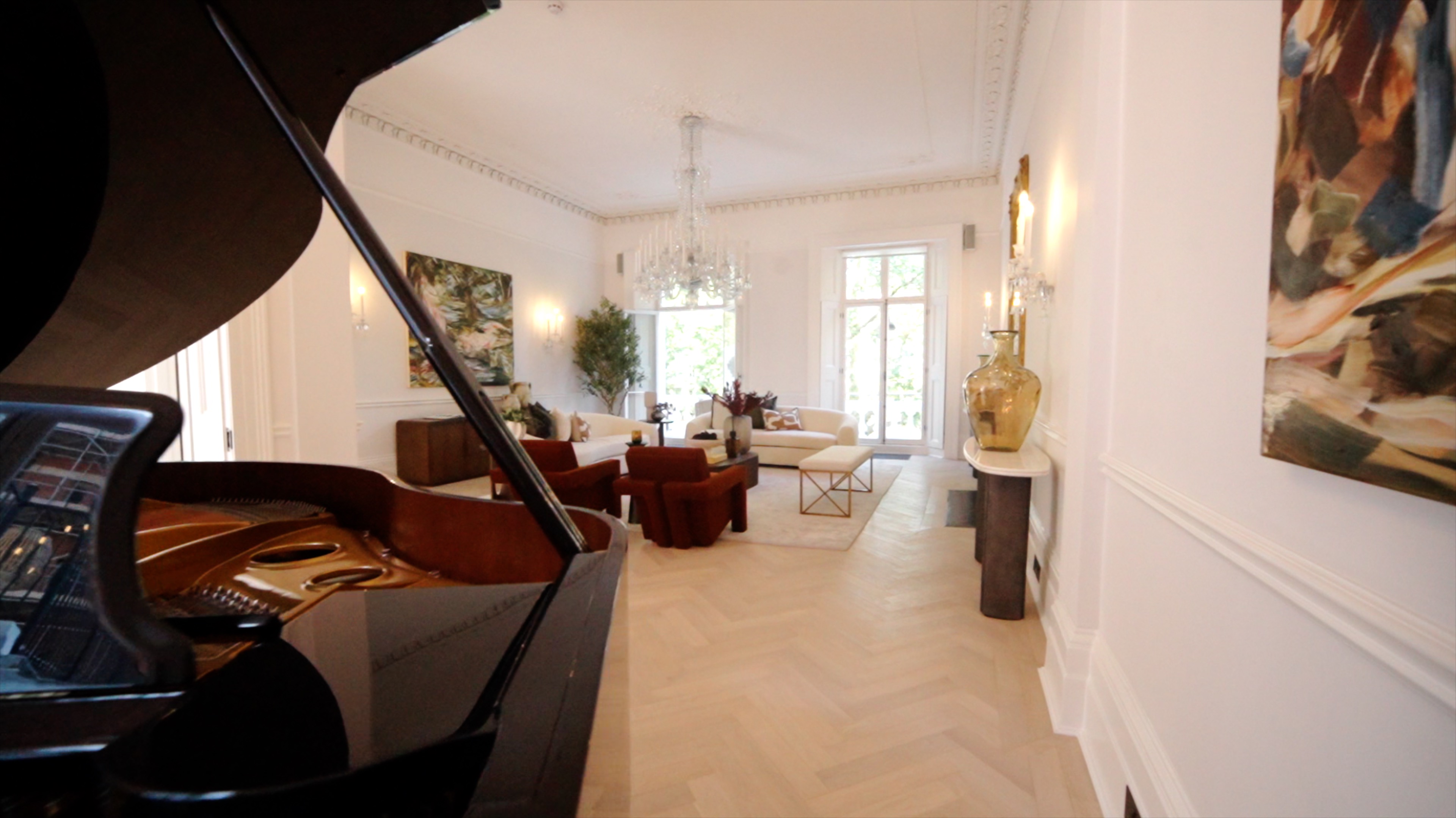 Bright living room with grand piano, crystal chandelier, white couches, and red armchairs on light wooden floor.
