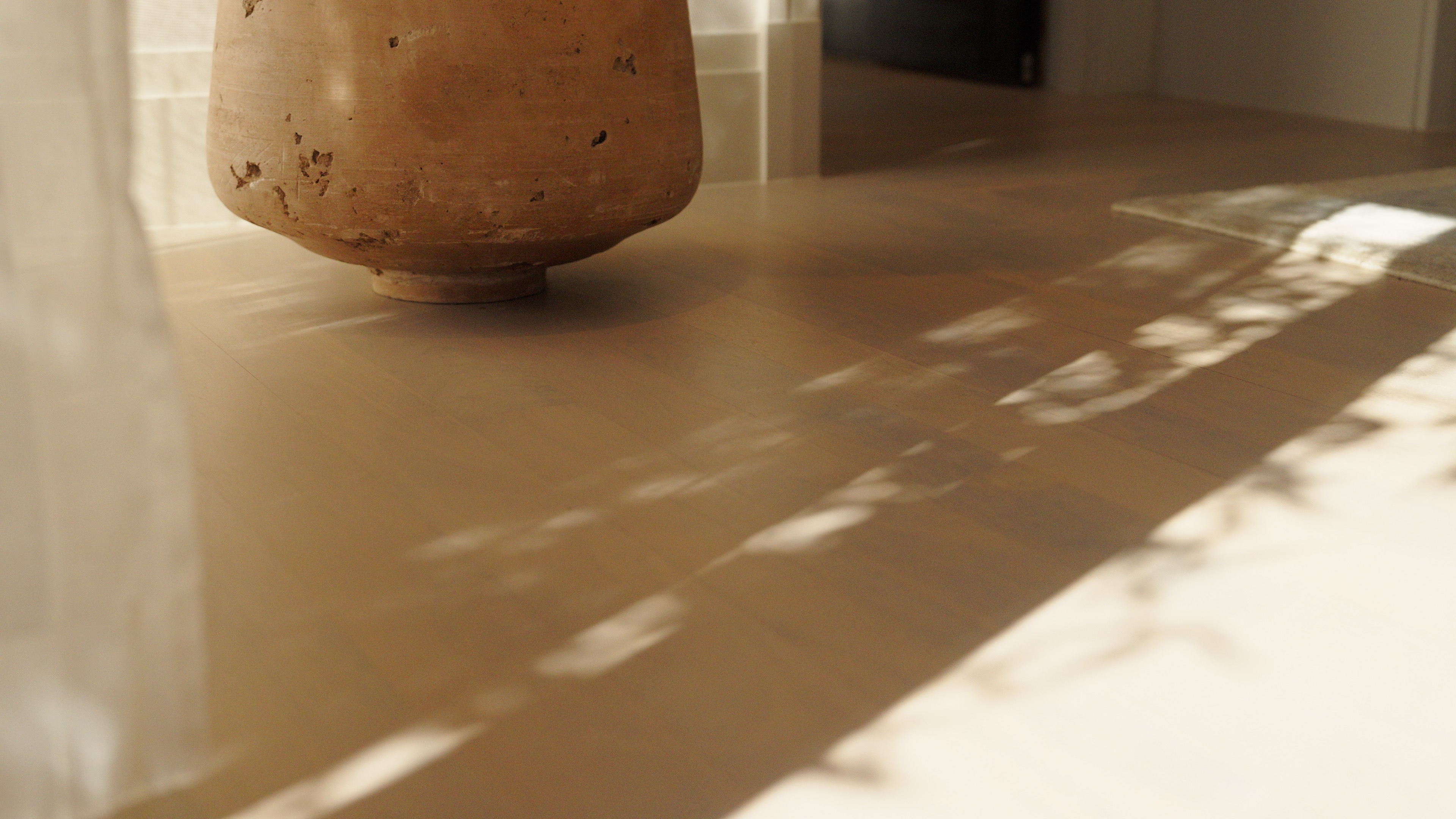 Large rustic clay pot casting shadow on a wooden floor with sunlight and curtain reflections.