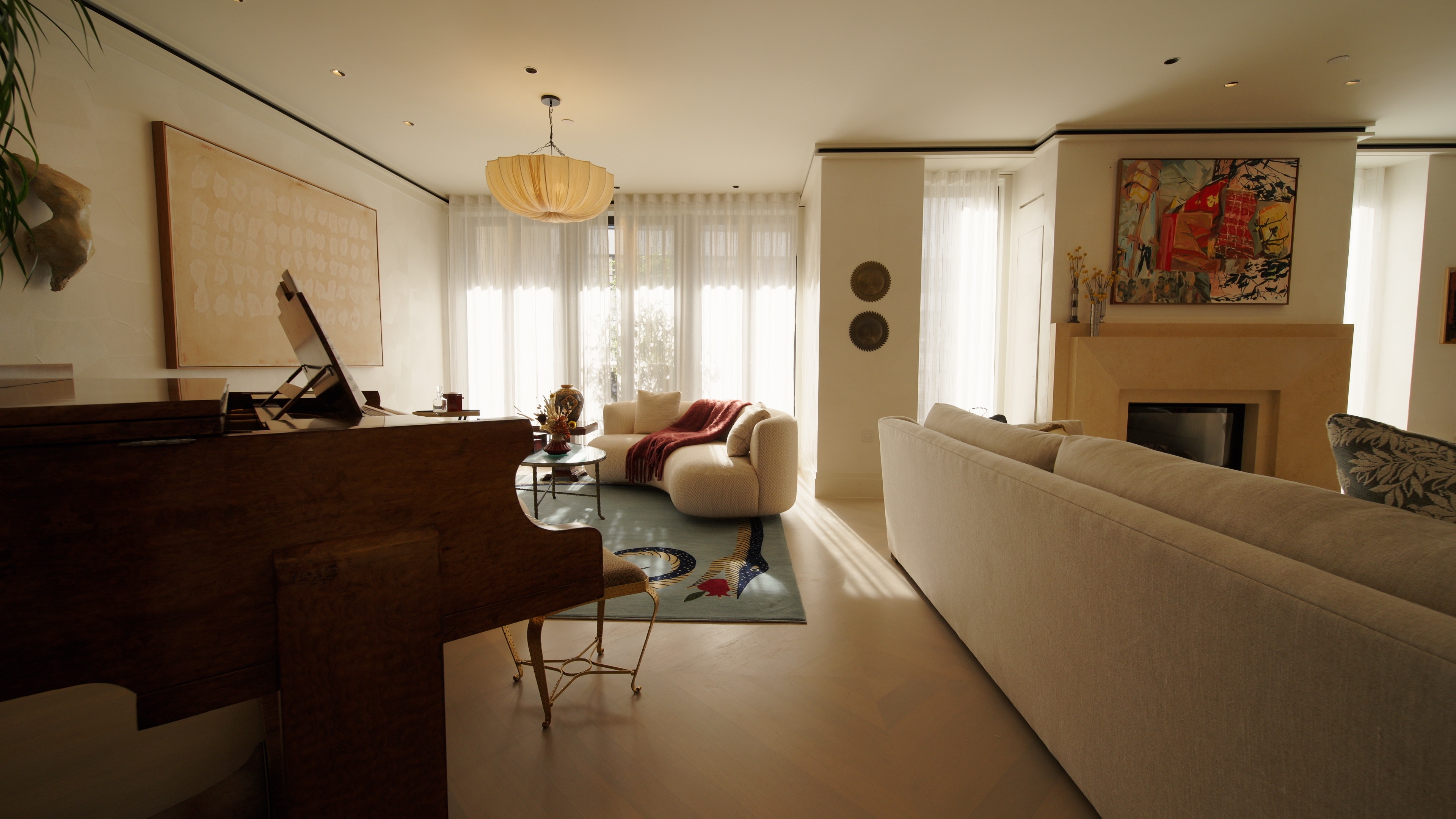 Living room with large beige sofas, a wooden piano, sheer curtains, and modern artwork above a fireplace.