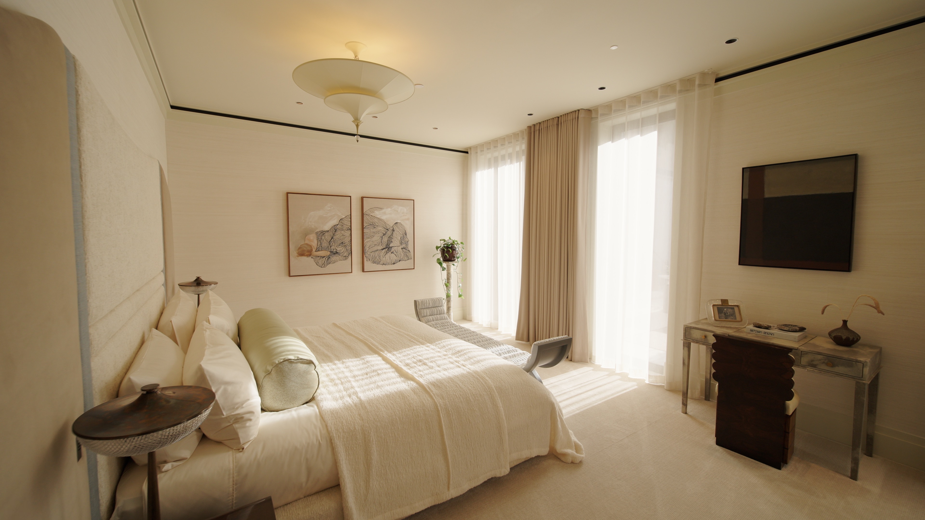 Bright bedroom with beige walls, large bed, chaise lounge, desk with chair, and sheer curtains on tall windows.