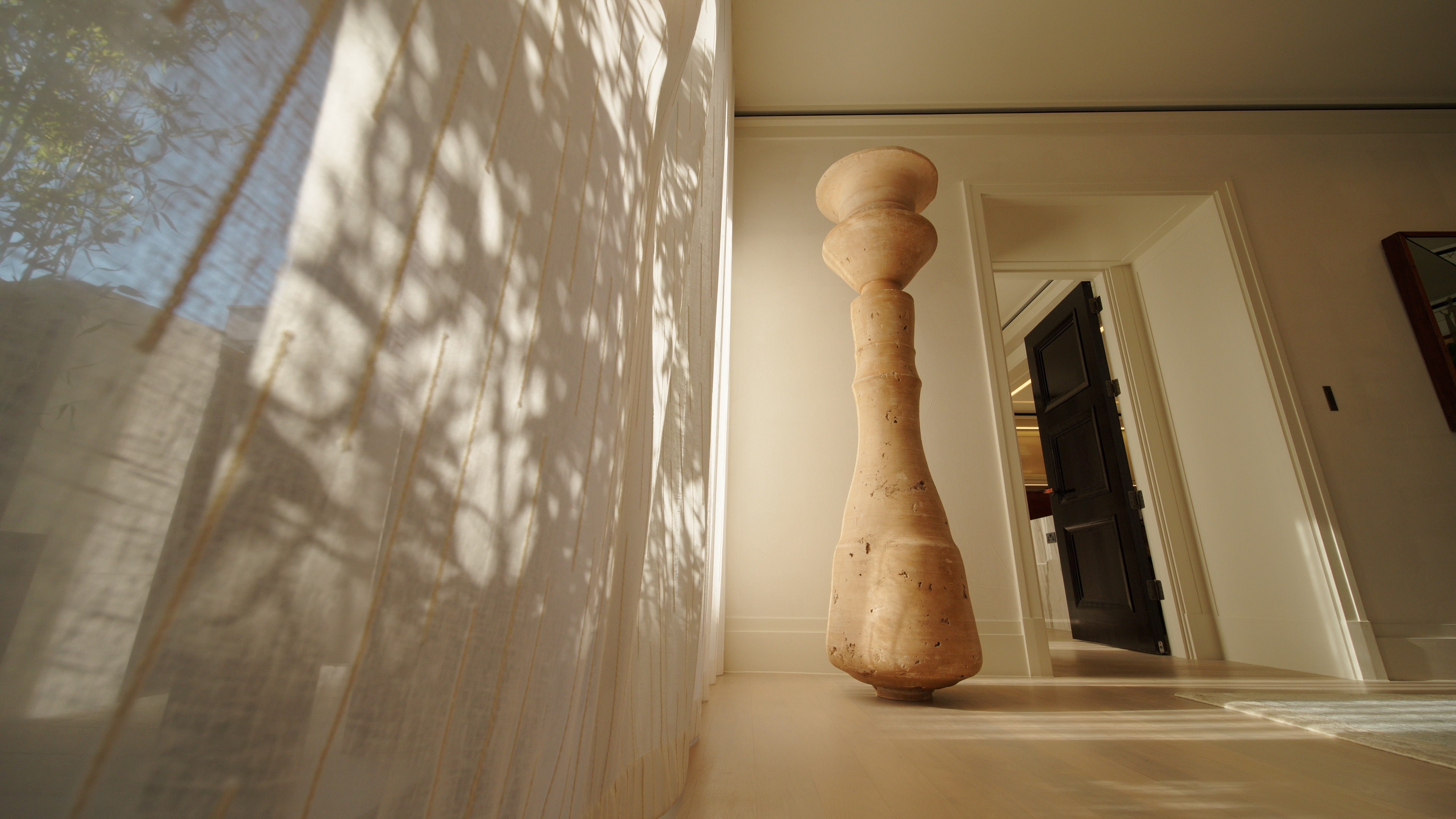 Tall, carved wooden sculpture stands on a light wooden floor near a sheer curtain and open door.