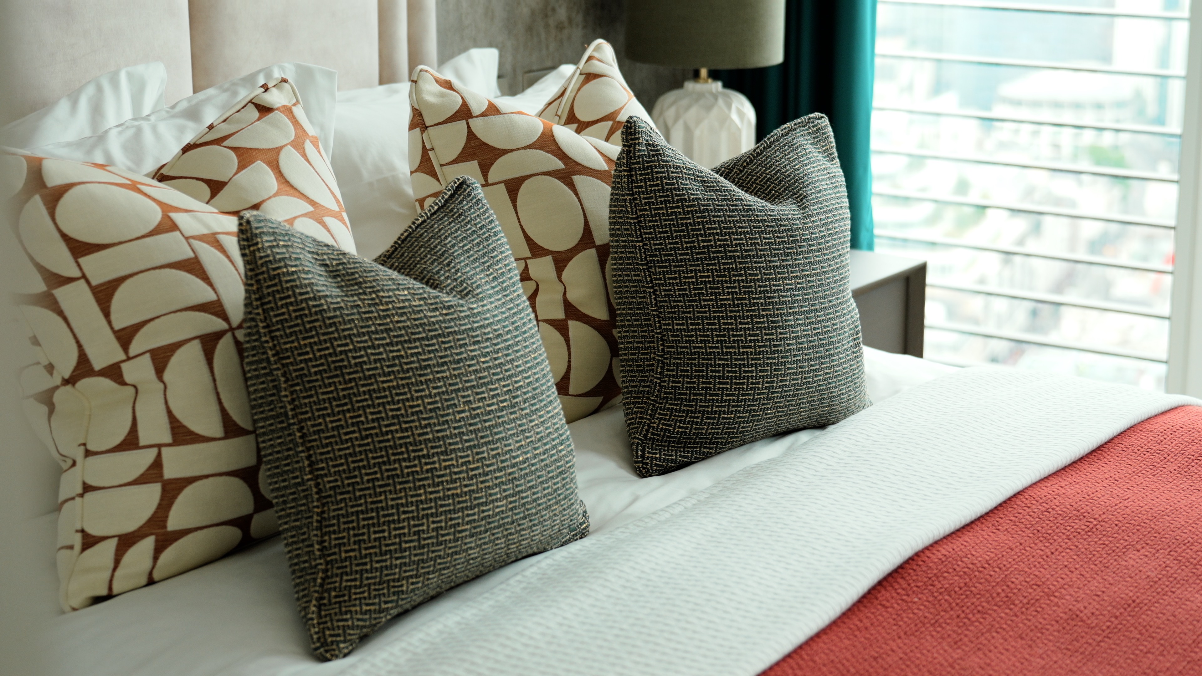 Four decorative pillows with geometric patterns on a white bed with a red blanket near a window.