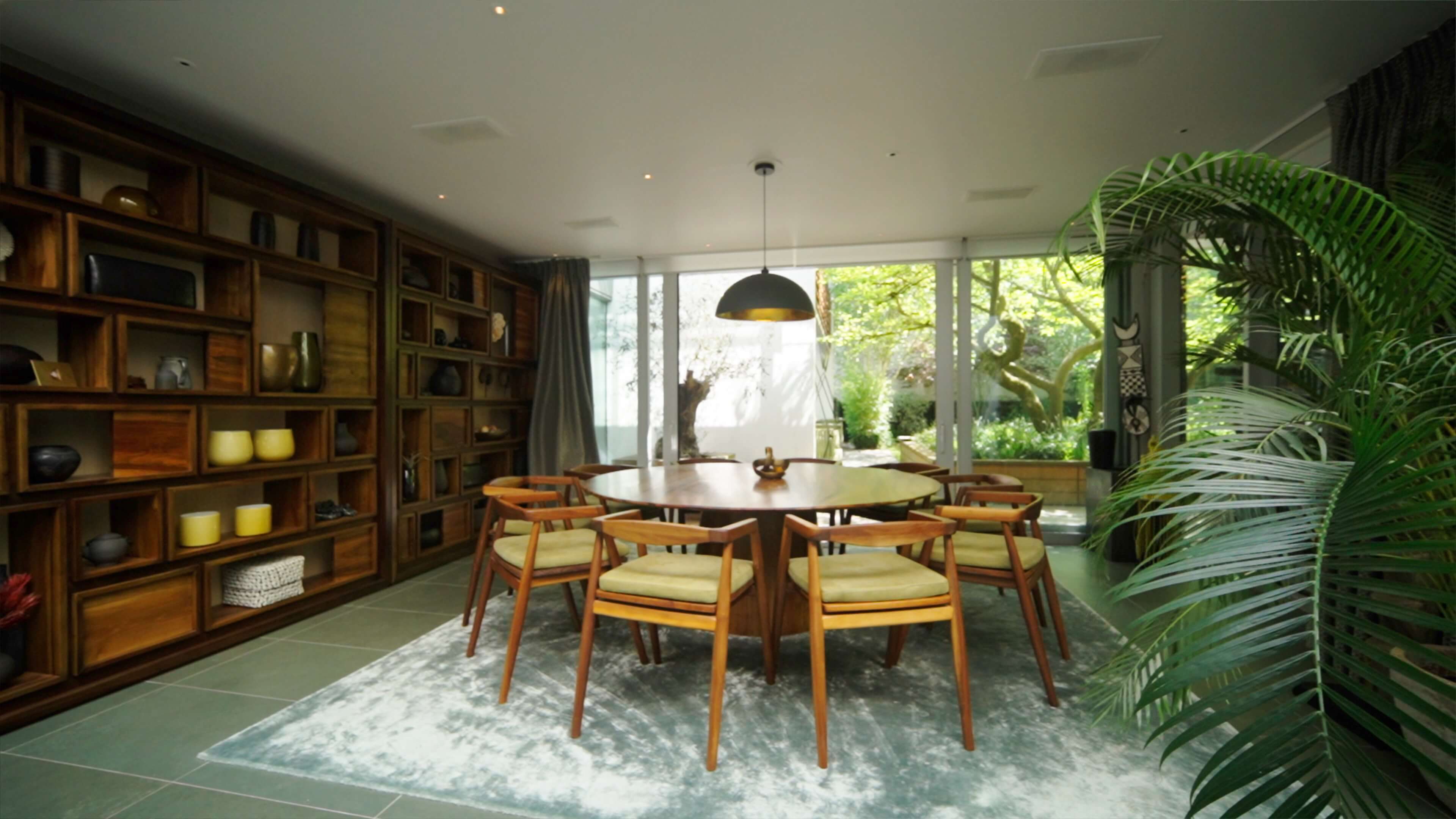 Dining room with round wooden table, eight chairs, large shelves, and glass wall opening to garden.