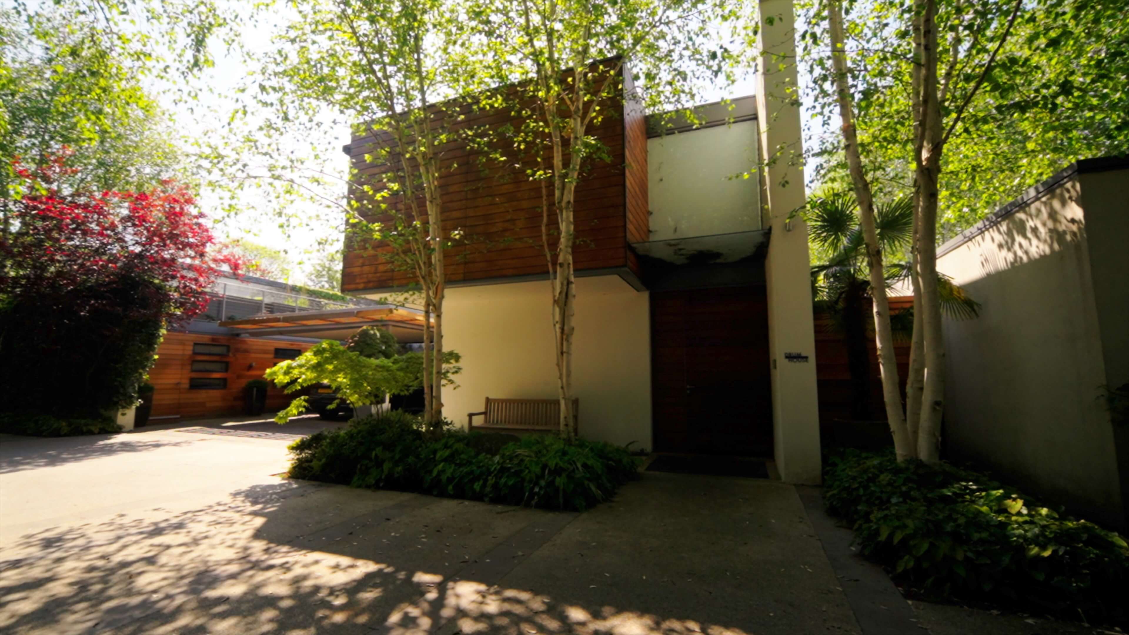 Modern house entrance with trees, wooden siding, bench, and shaded driveway.