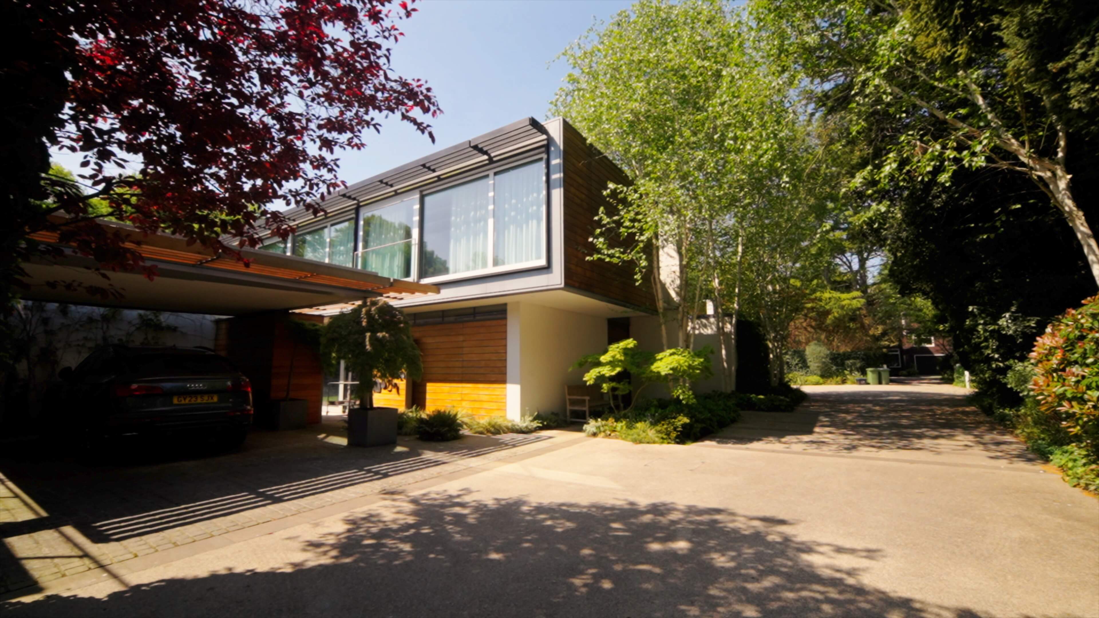 Modern house with large windows, wooden panels, carport with black car, and leafy trees and shrubs nearby.