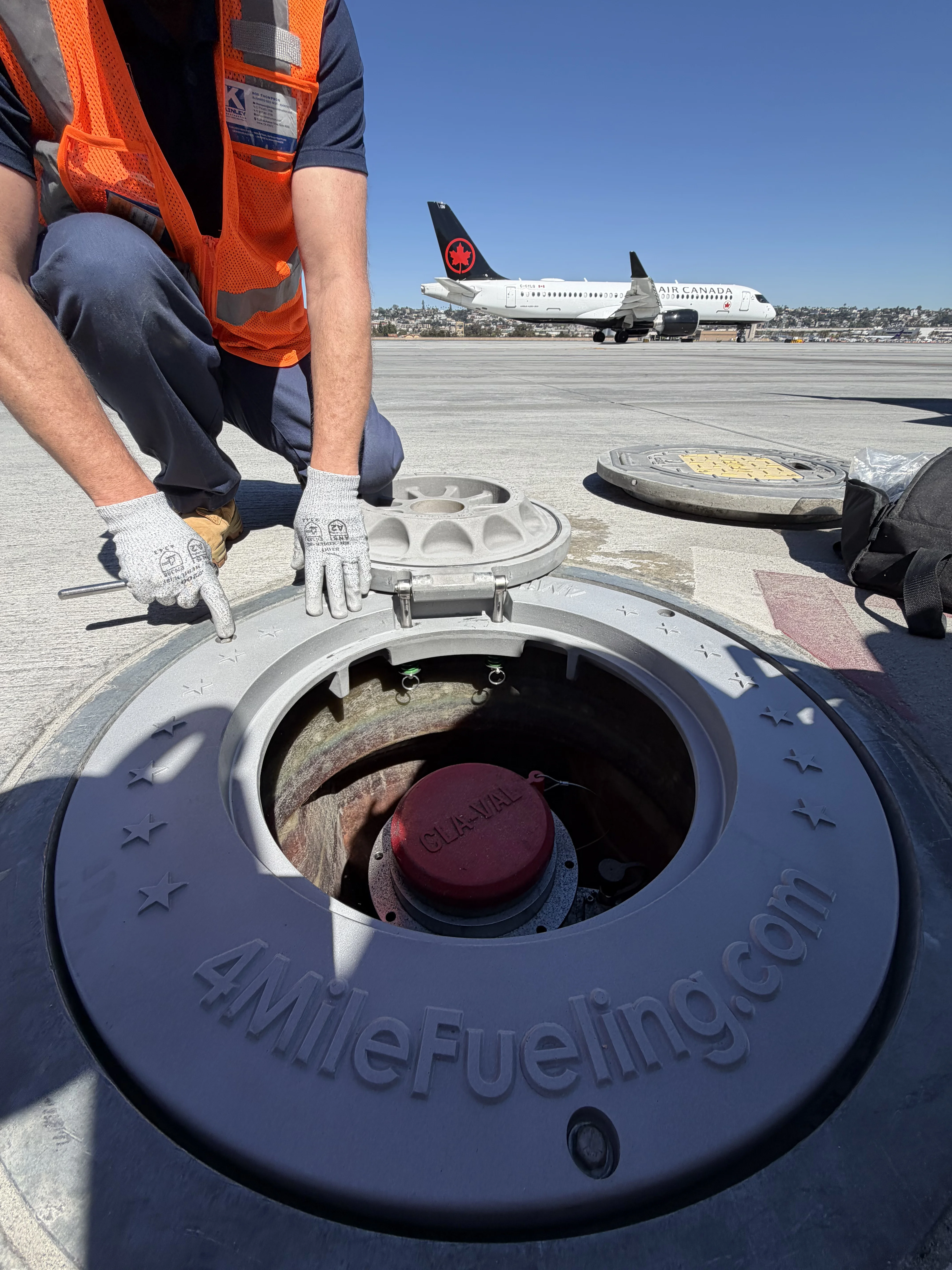 A worker wearing an orange safety vest and gloves opens a circular aircraft fueling pit labeled '4MileFueling.com' on an airport tarmac. An Air Canada airplane is visible taxiing in the background under a clear blue sky.