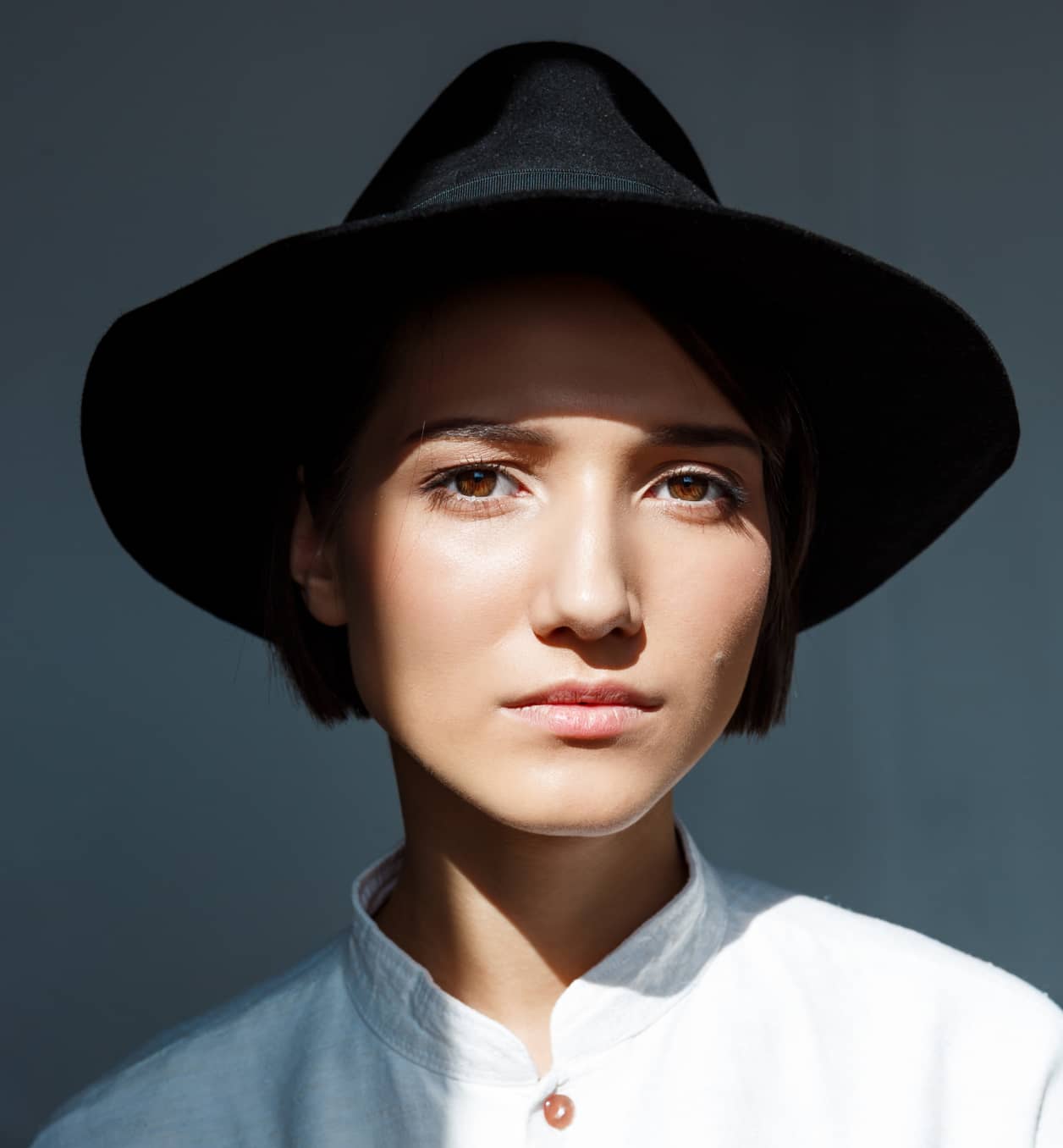 Portrait of a woman with brown eyes wearing a black wide-brimmed hat and a white shirt, lit by sunlight.