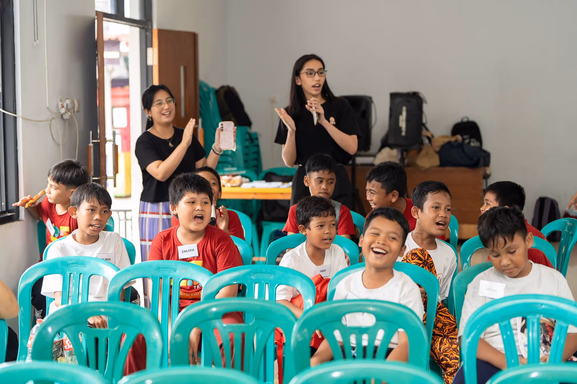 A group of children sitting on green plastic chairs, some laughing and talking, with two women clapping and smiling behind them in a classroom setting.