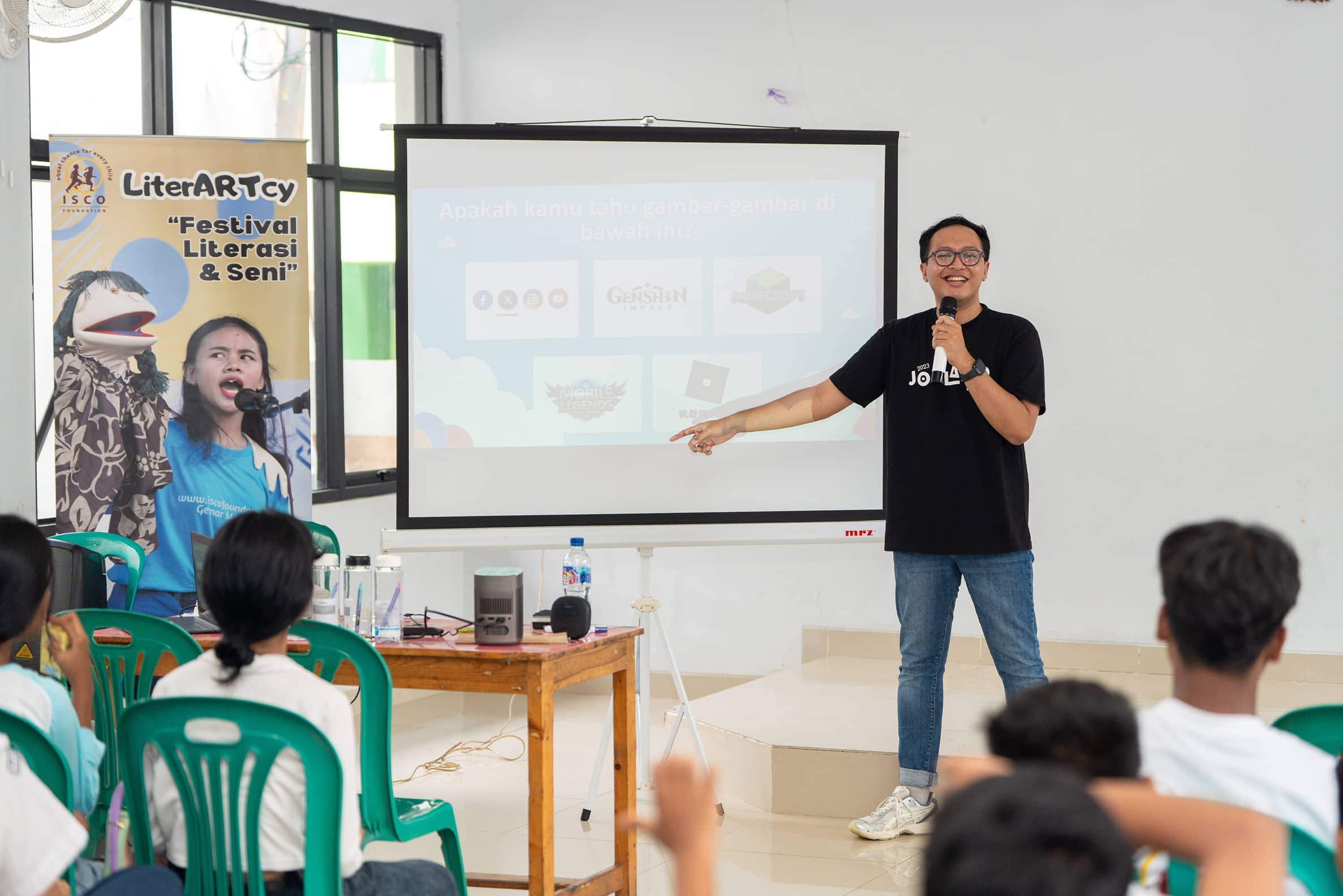 Man in glasses and black t-shirt holding a microphone and pointing at a projection screen during a literacy and arts festival presentation with seated audience.