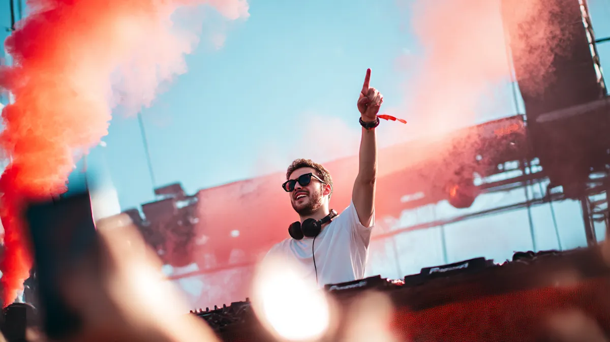 DJ wearing sunglasses and headphones performing at a music festival with red smoke behind.