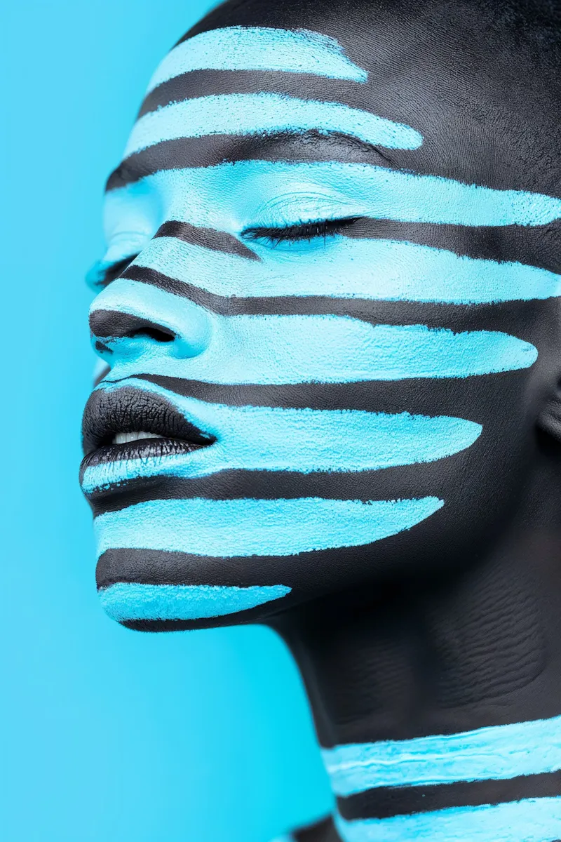 Close-up of a person with closed eyes and blue and black striped face paint on a blue background.