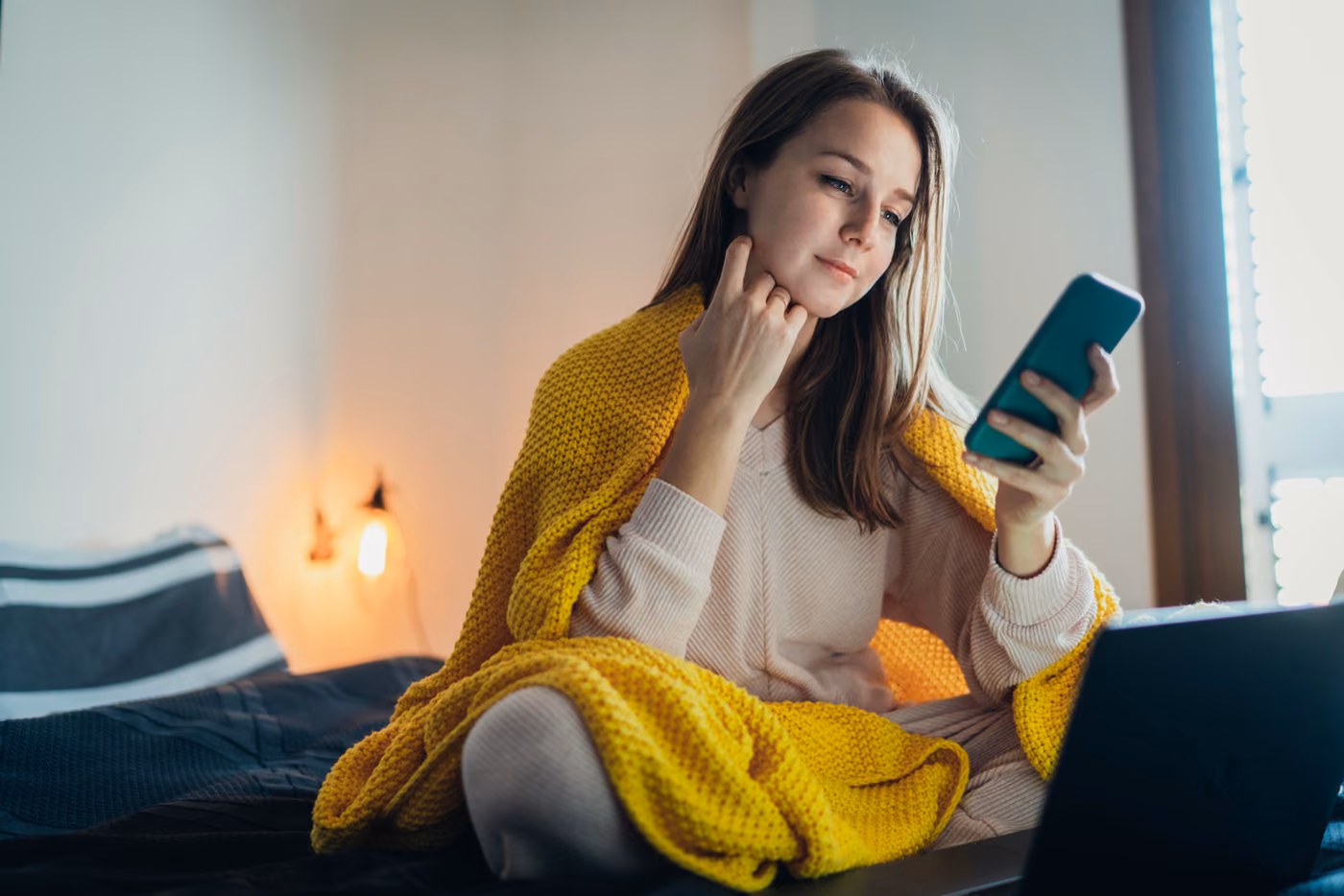 a woman sits on her bed wrapped in a yellow blanket looking at her phone