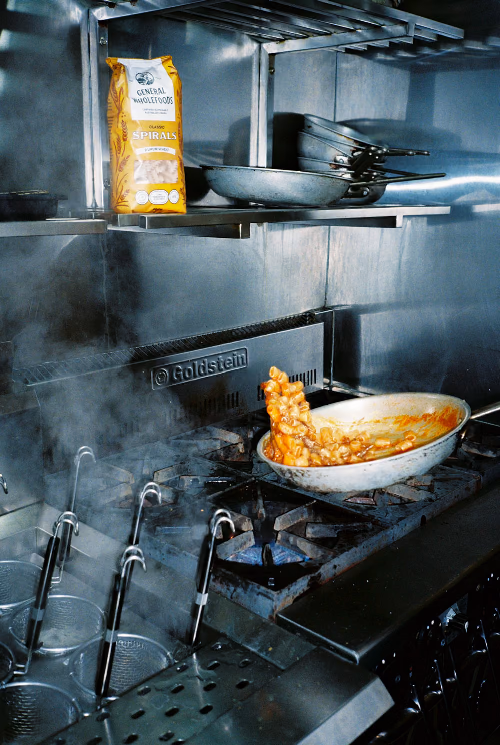 General Wholefoods pasta cooking in sauce flipped mid-air in a pan on a commercial gas stove with steam rising.